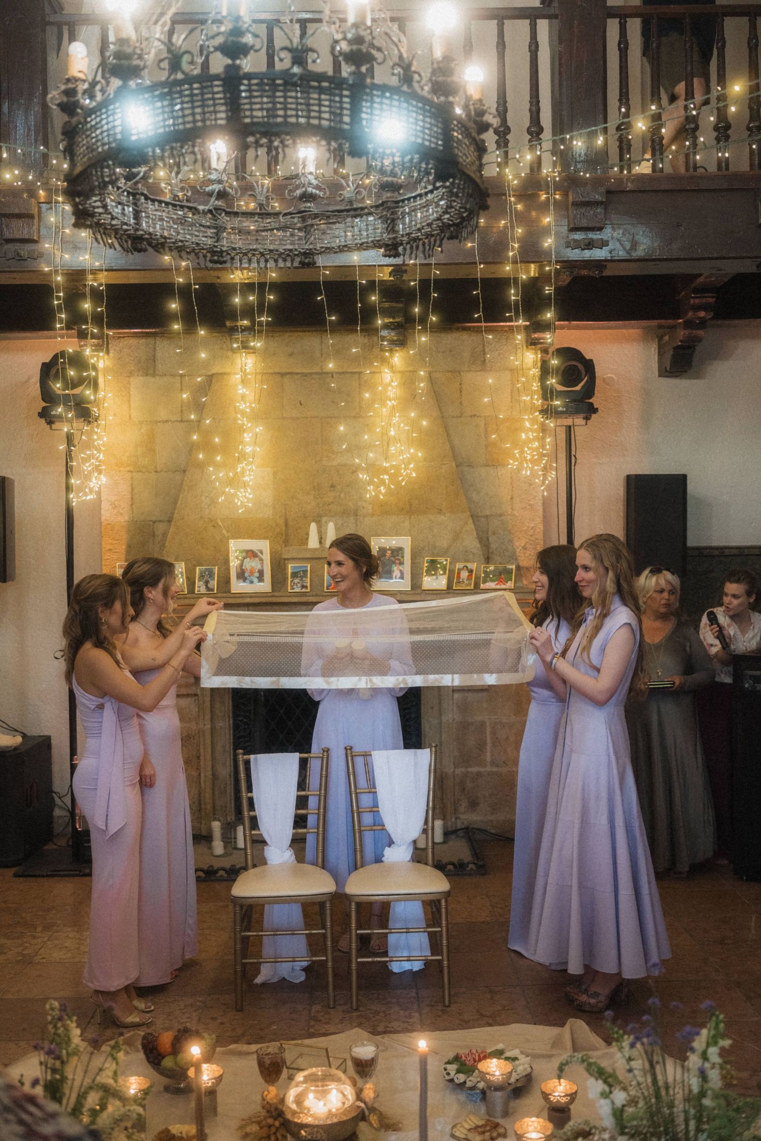 Bridesmaids in purple dresses holding a veil over a bride at a candlelit wedding.