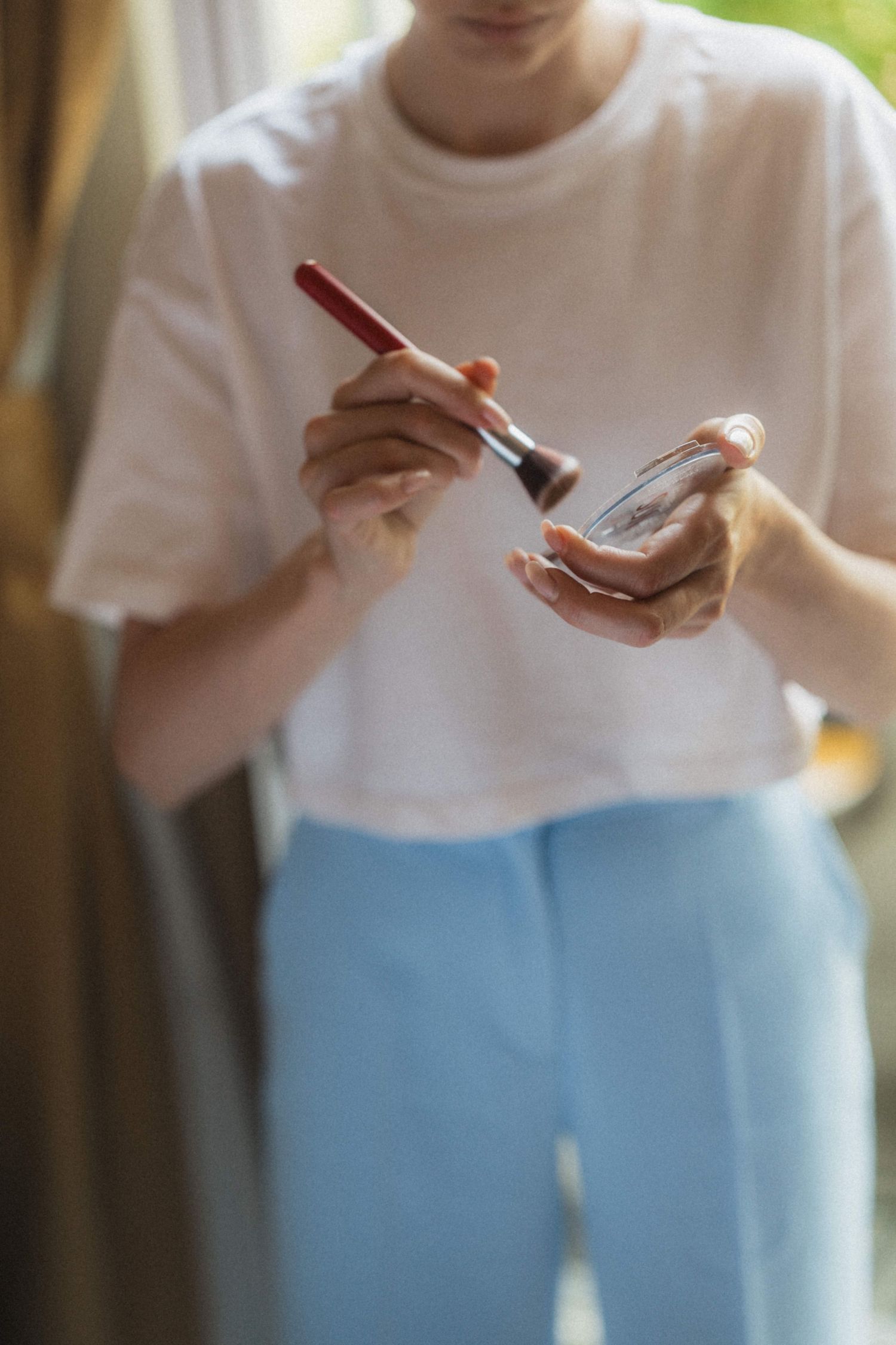 Woman in white tee and blue pants holding a makeup brush and compact mirror while applying cosmetics.