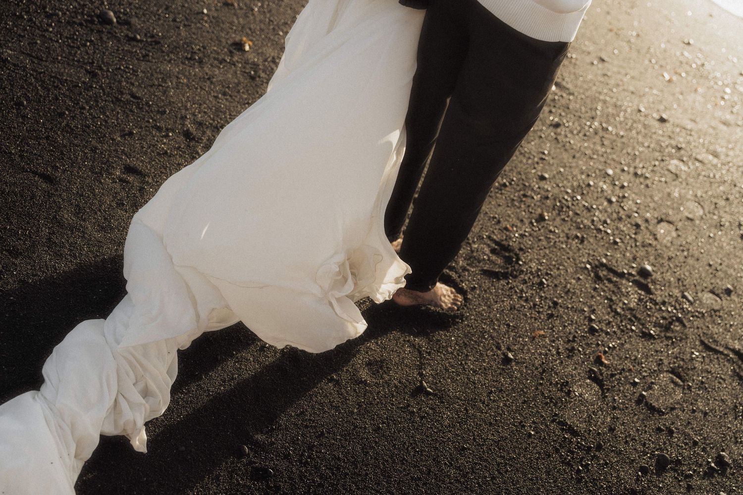 Close-up of wedding dress train and groom shoes on dark volcanic black sand beach.