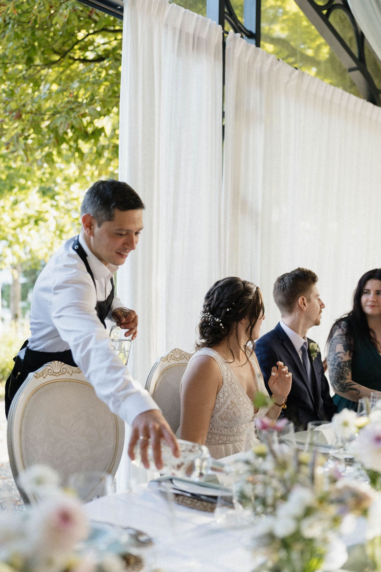 Waiter serving bride and groom at elegant outdoor wedding reception with floral centerpieces.