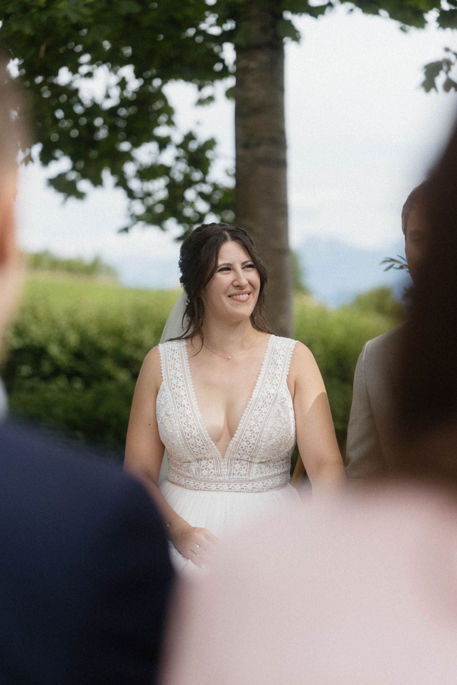 Smiling bride in a white lace V-neck gown stands outdoors during a wedding ceremony with greenery in the background.
