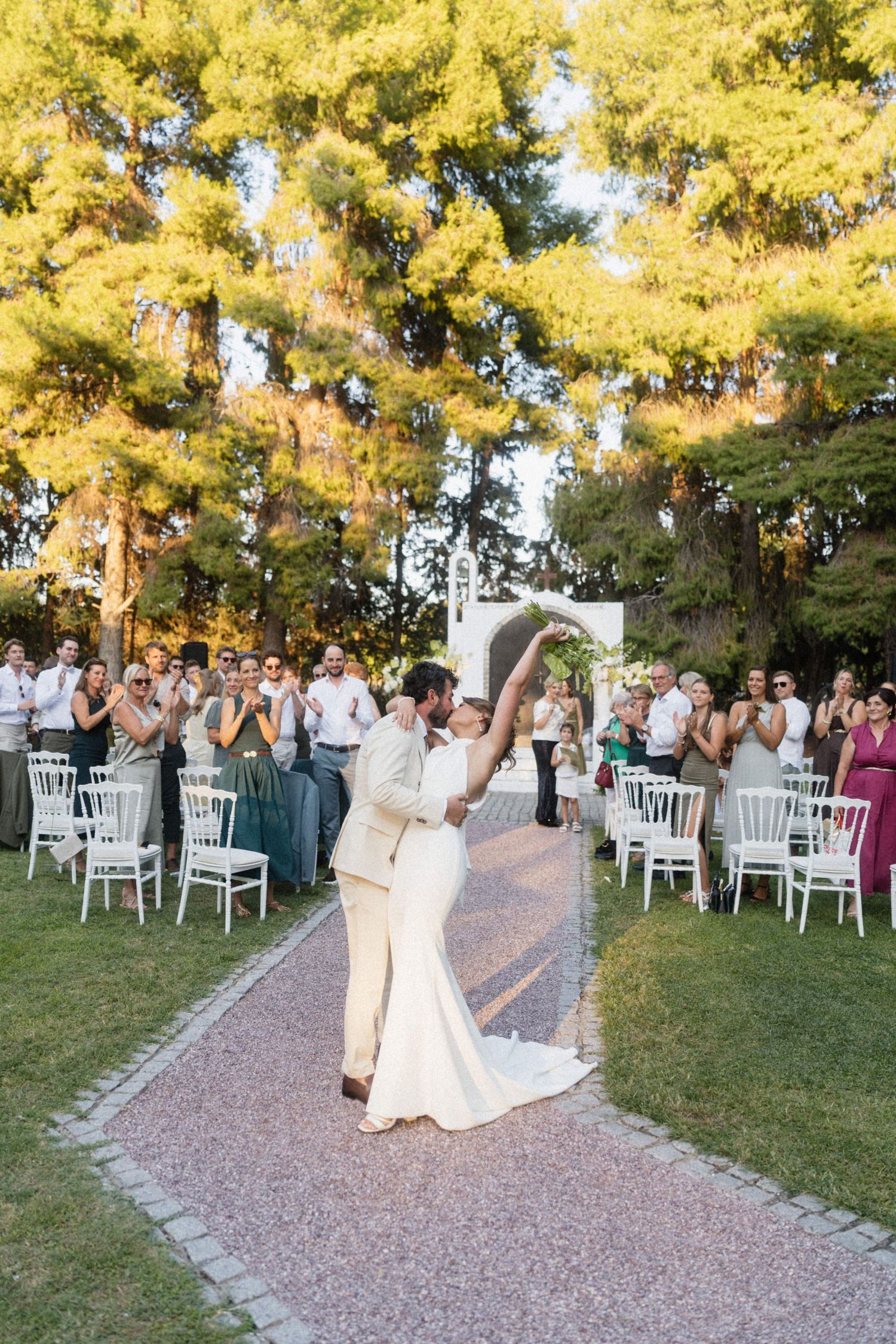 Newlyweds celebrate joyfully on garden aisle as guests applaud, with tall trees glowing in warm sunset light behind them.