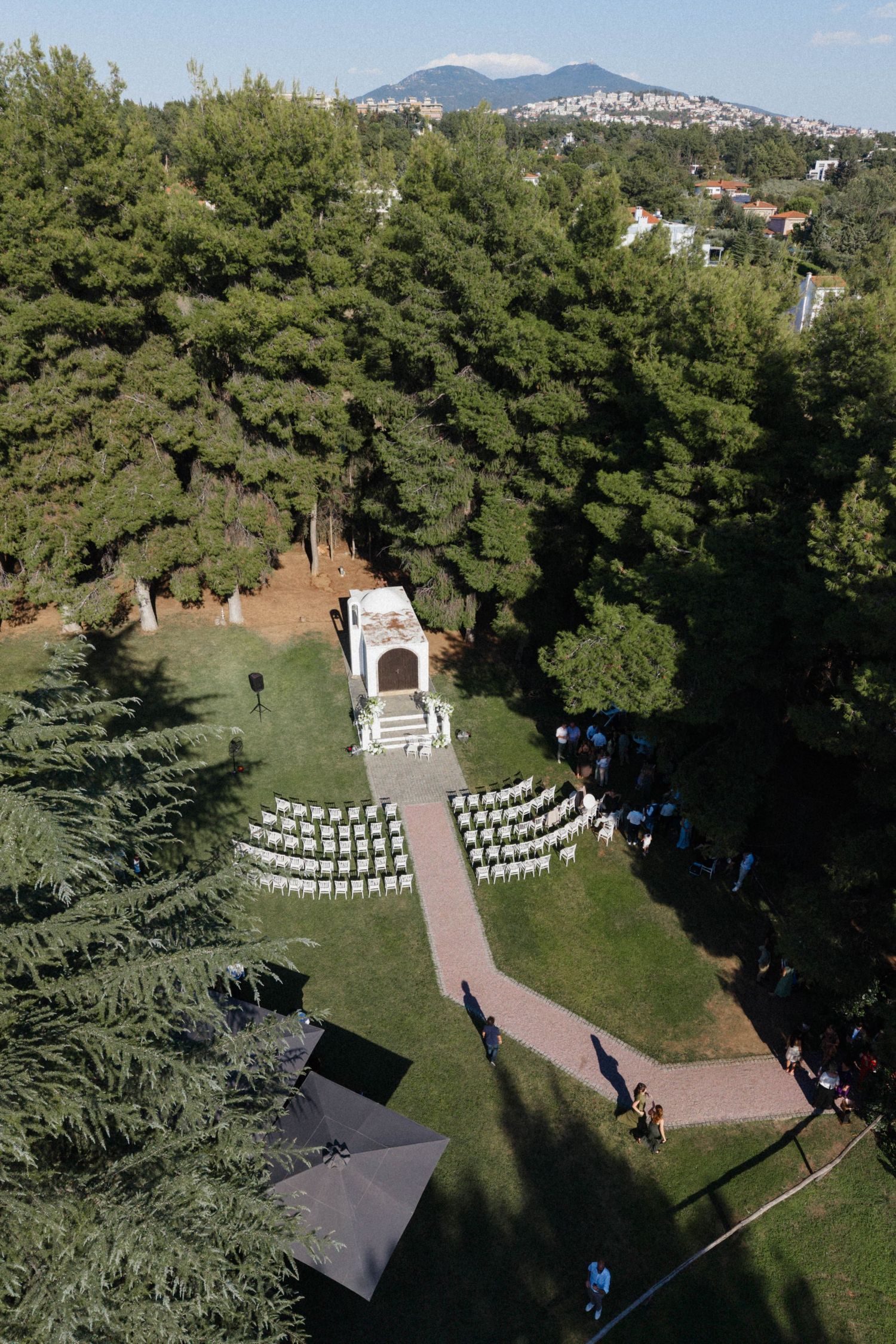 Aerial view of an outdoor wedding ceremony setup with white chairs in a lush green garden near a historic chapel.