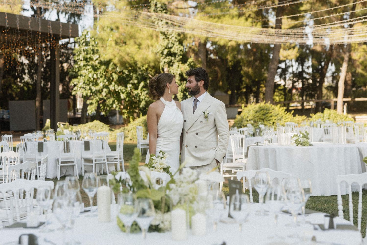 Bride and groom share an intimate moment surrounded by elegantly set white reception tables in a sunlit garden.