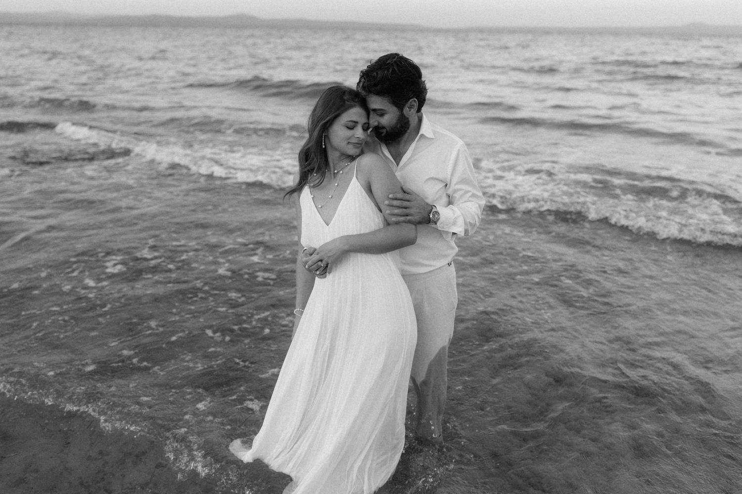 Black and white photo of a romantic couple embracing on a beach, waves surrounding them, woman in flowing white dress.