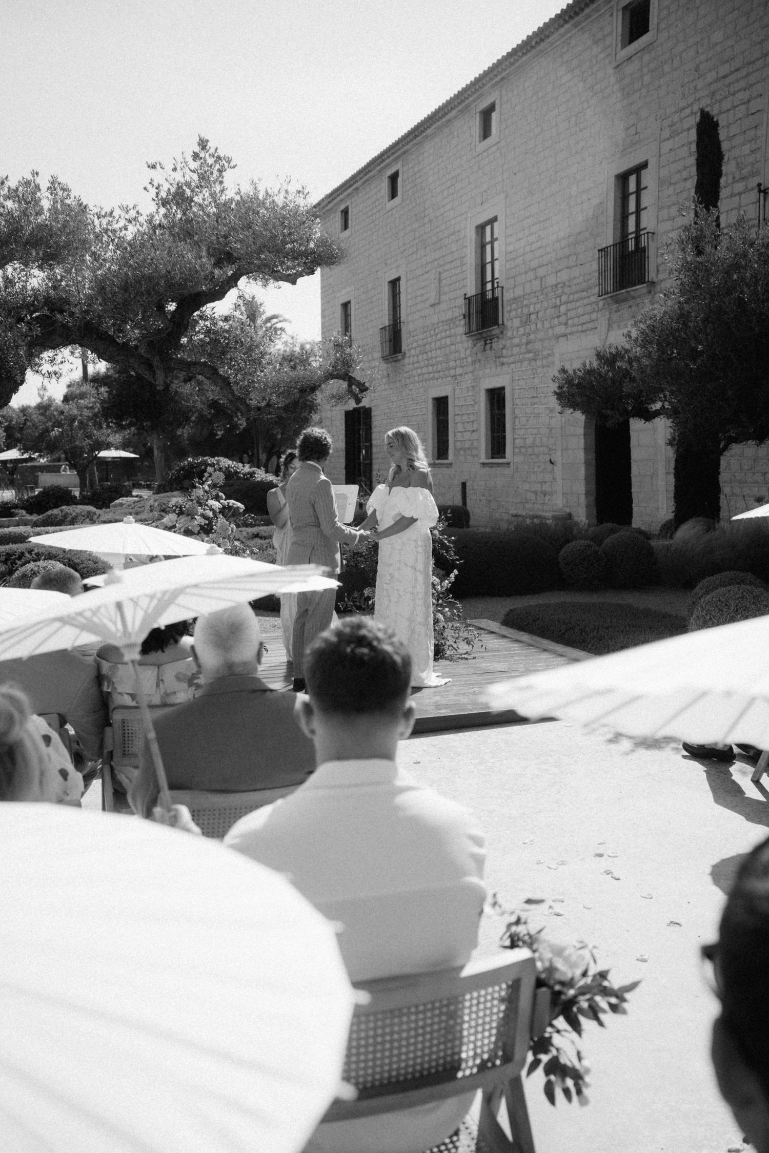 Black and white photo of outdoor wedding ceremony with guests seated under parasols at stone estate.