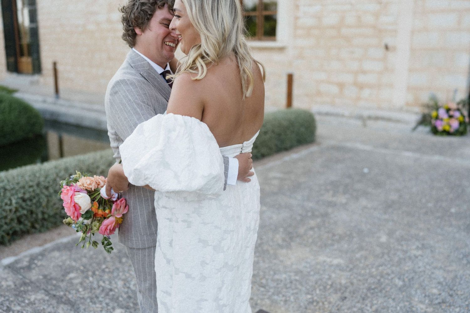 Bride in white off-shoulder gown holds pink bouquet while groom in gray suit embraces her outdoors.