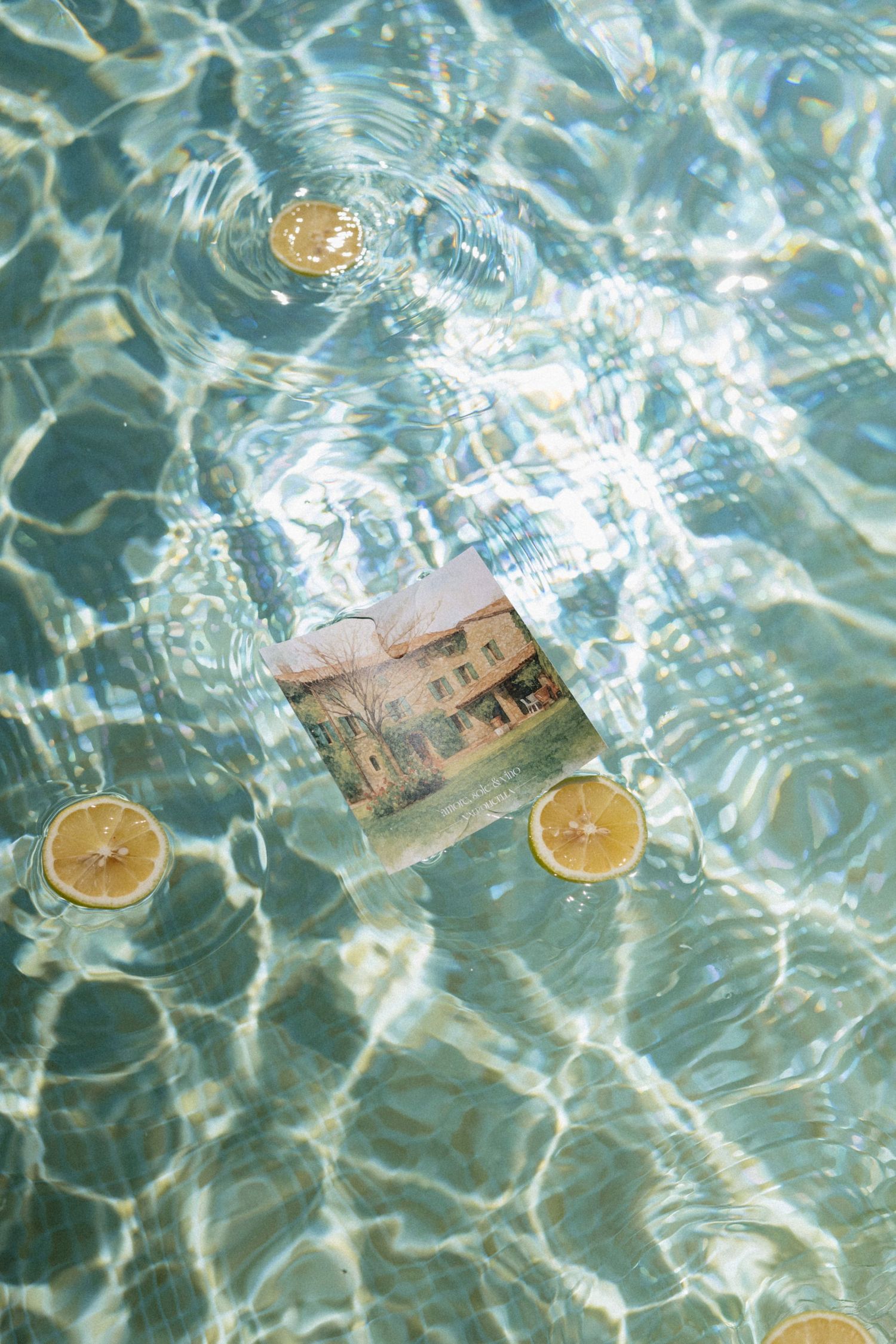 Lemon slices and a vintage postcard floating in clear turquoise pool water with rippling light reflections.