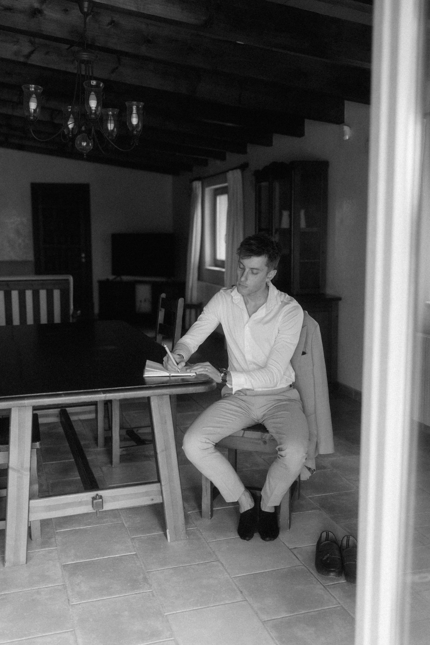 Black and white photo of a man in white shirt writing at a wooden table in a rustic room.