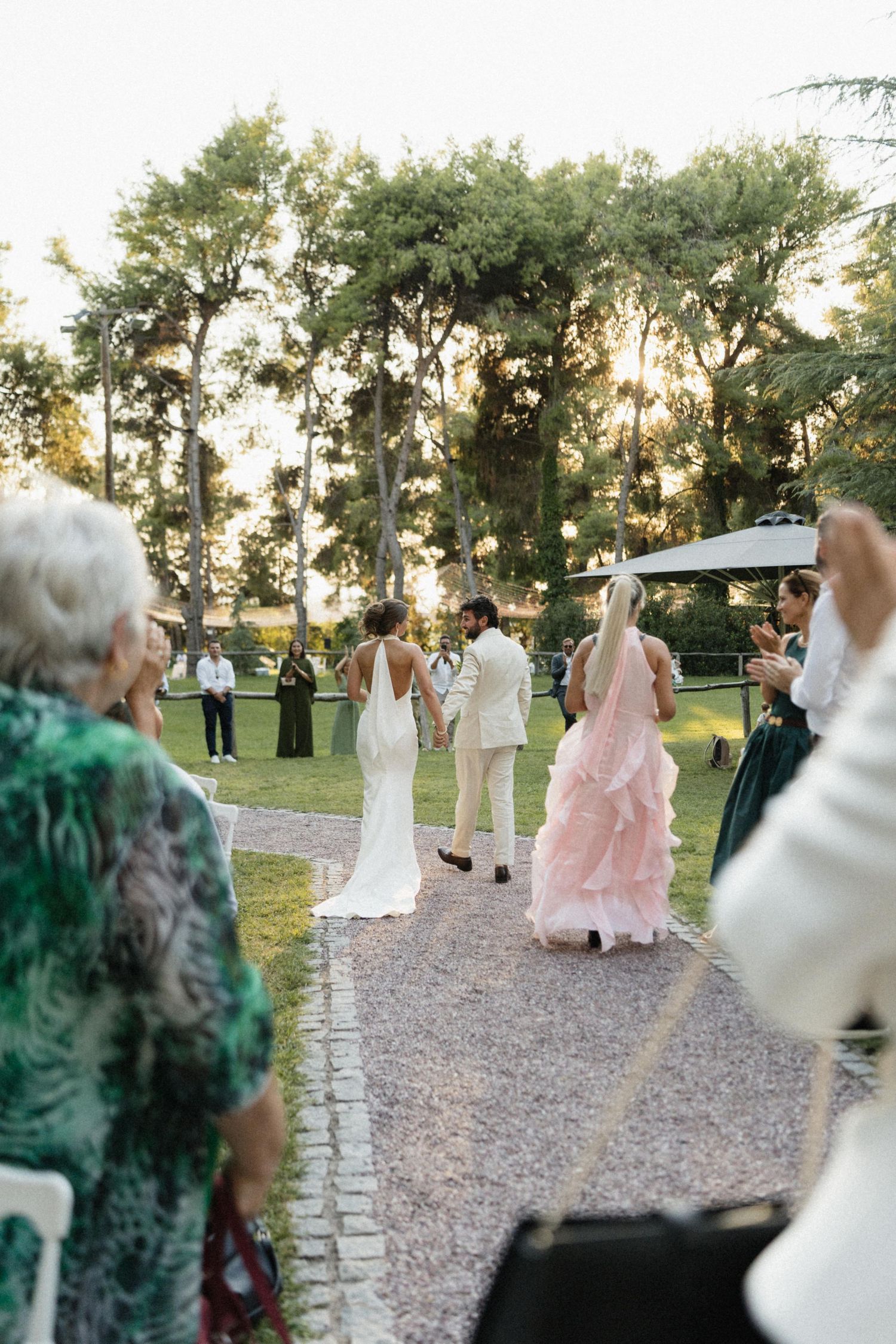 Bride in white gown and groom in cream suit walk down aisle with guests applauding at golden hour outdoor wedding.