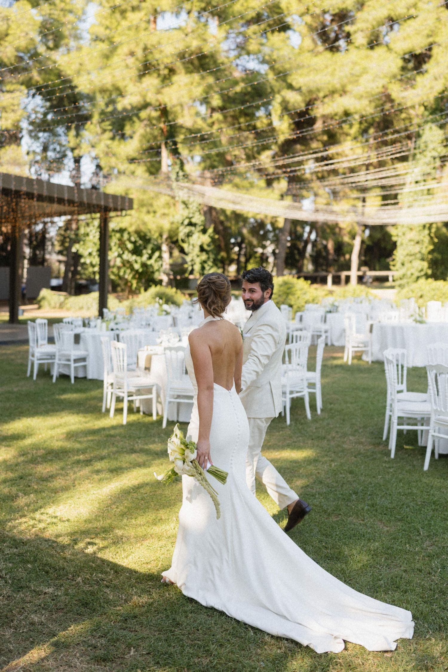 Bride and groom walking through an elegant outdoor wedding reception with white chairs and tables on a green lawn.