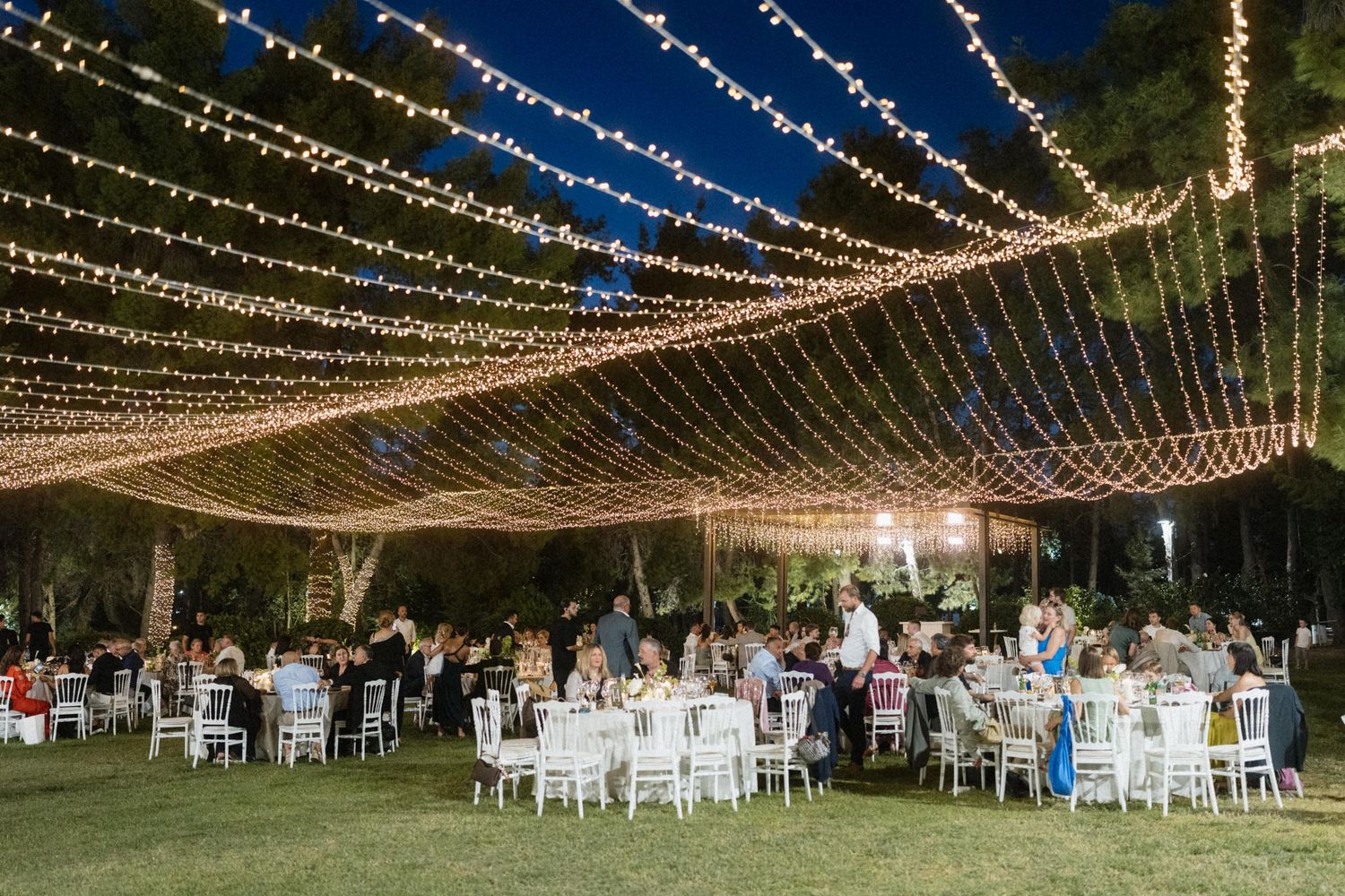 Outdoor evening wedding reception with string lights canopy over white tables and chairs on green lawn.
