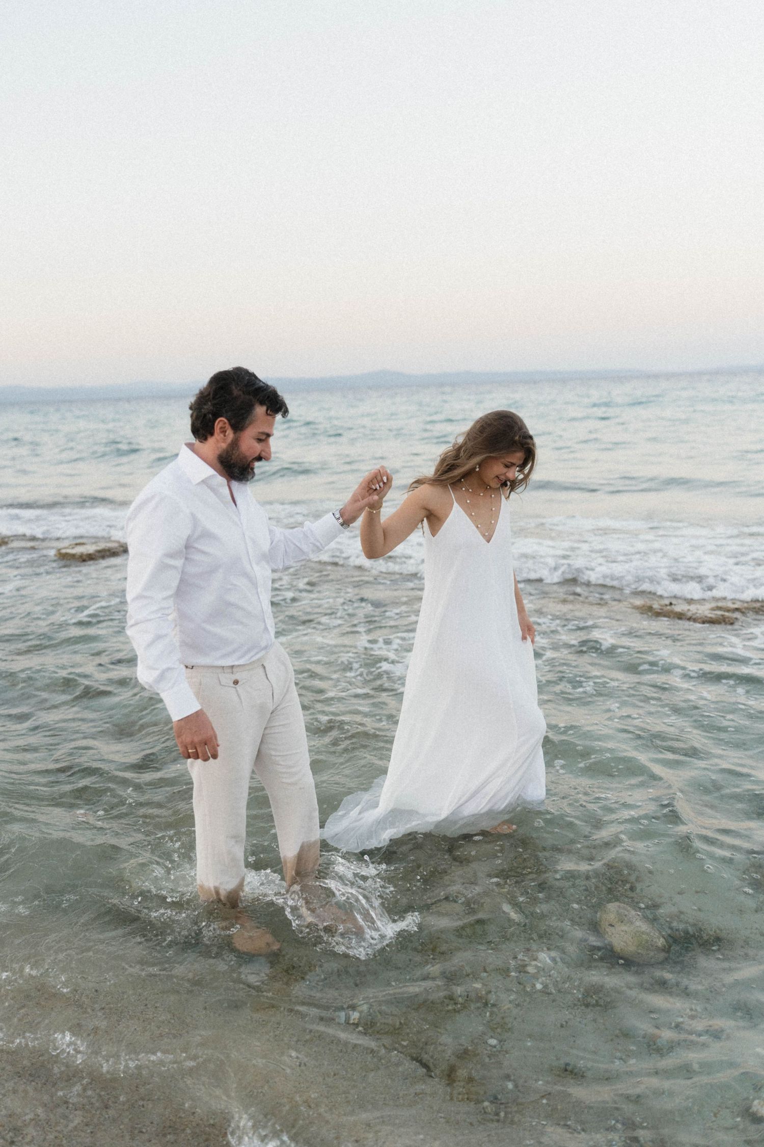 Groom in white shirt helps bride lift her flowing white dress while wading together through shallow ocean waves.