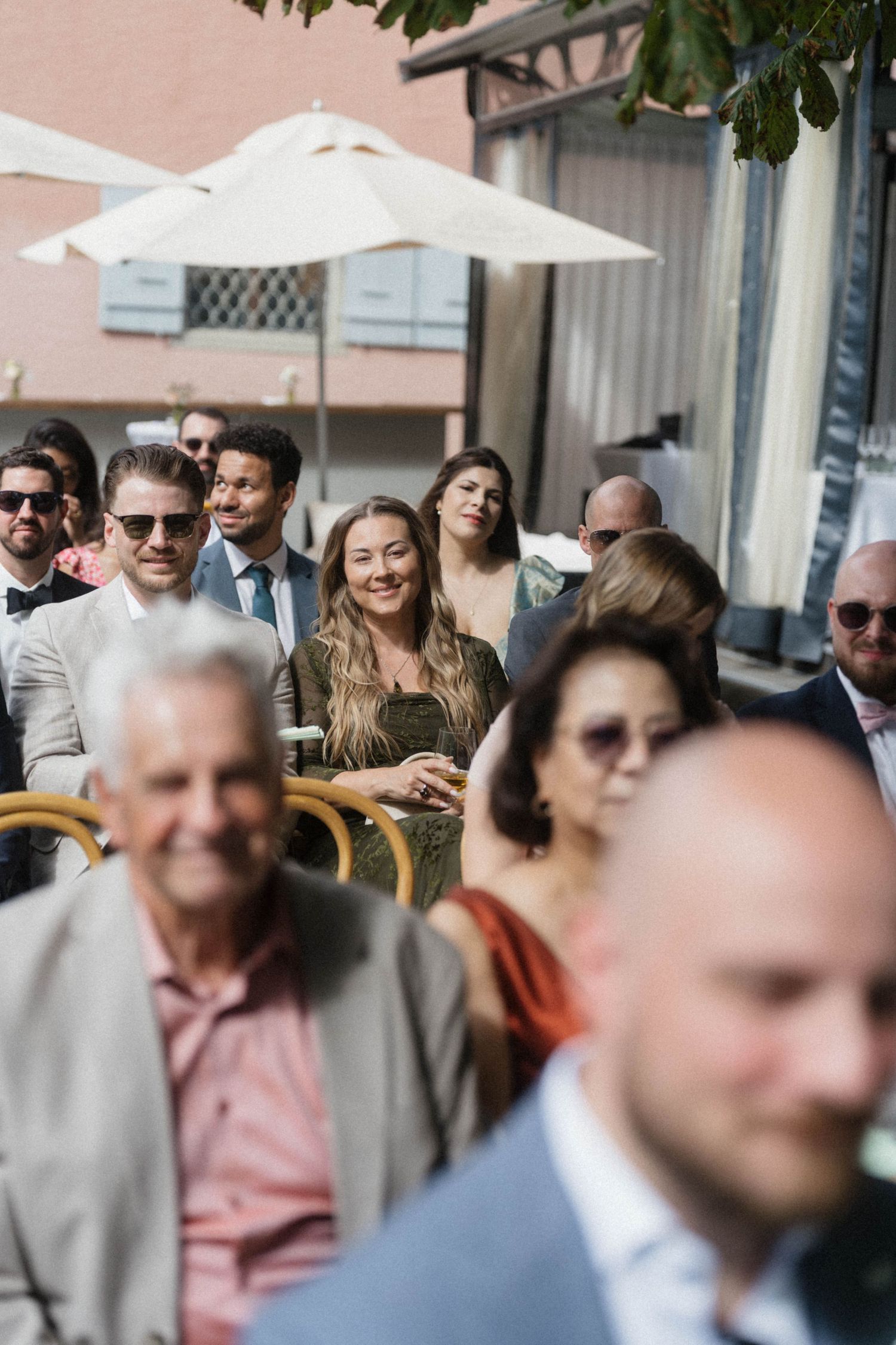 Wedding guests seated outdoors under white umbrellas, smiling woman in olive dress stands out.