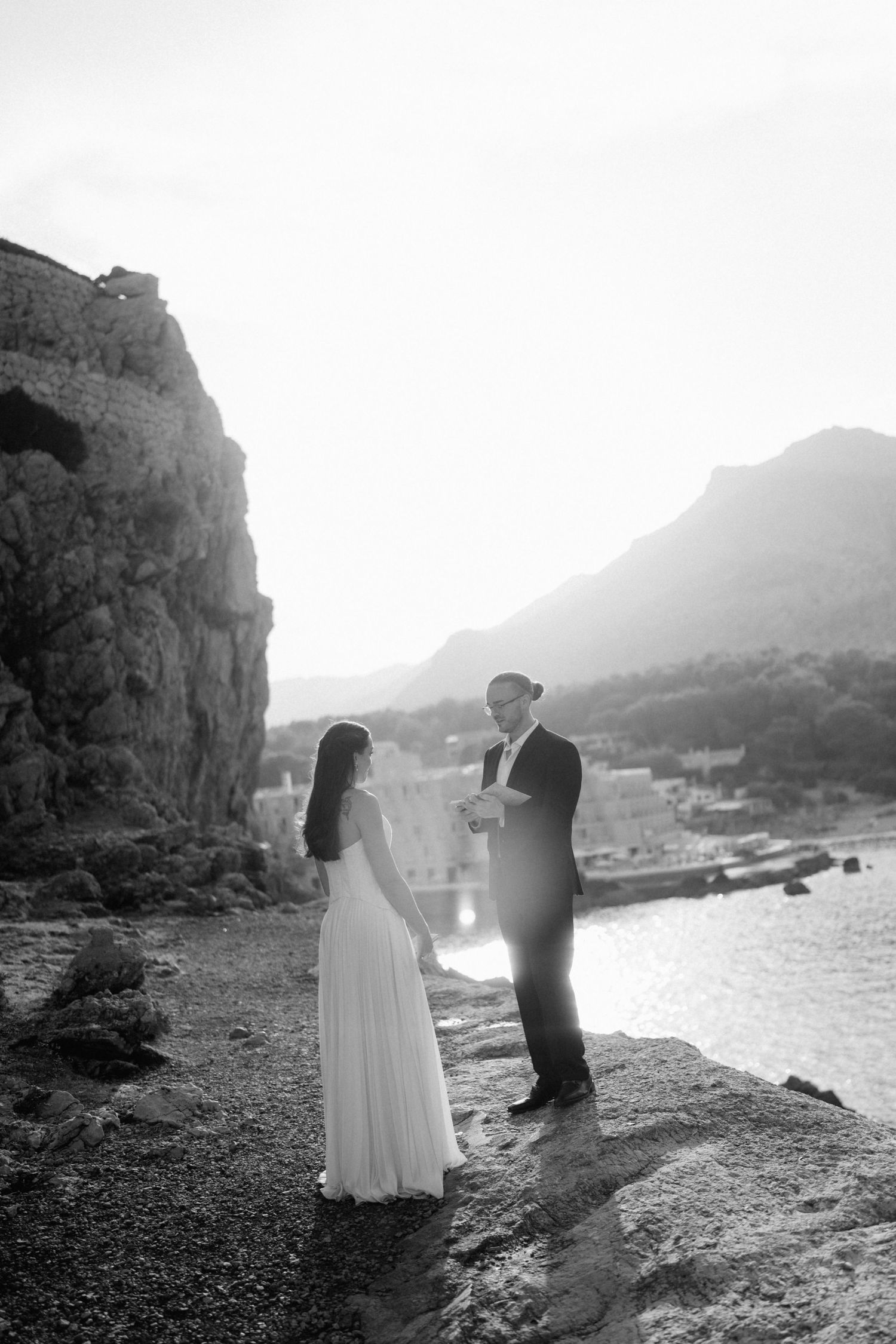 Black and white photo of a bride and groom exchanging vows on a rocky shore with a coastal village and mountains behind.