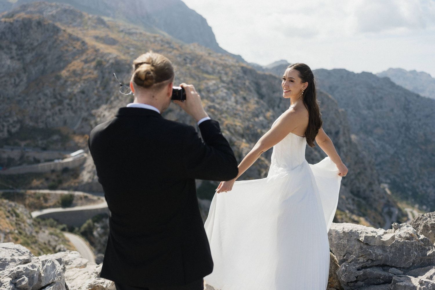 Bride in a flowing white wedding dress poses on a rocky mountain overlook while a groom in a black suit photographs her.