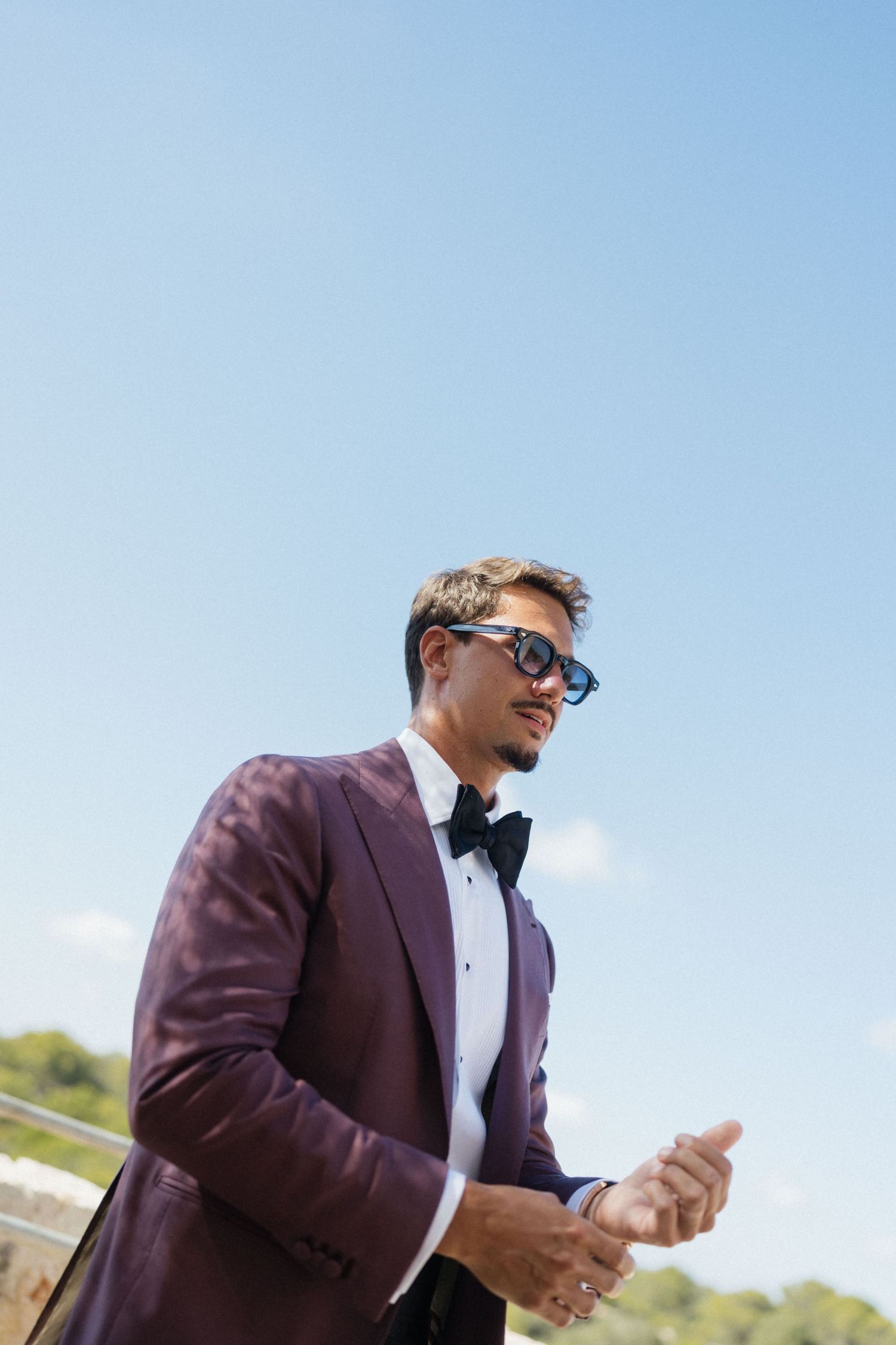 Stylish groom in a burgundy blazer, black bow tie and blue sunglasses stands against a clear blue sky at an outdoor wedding.