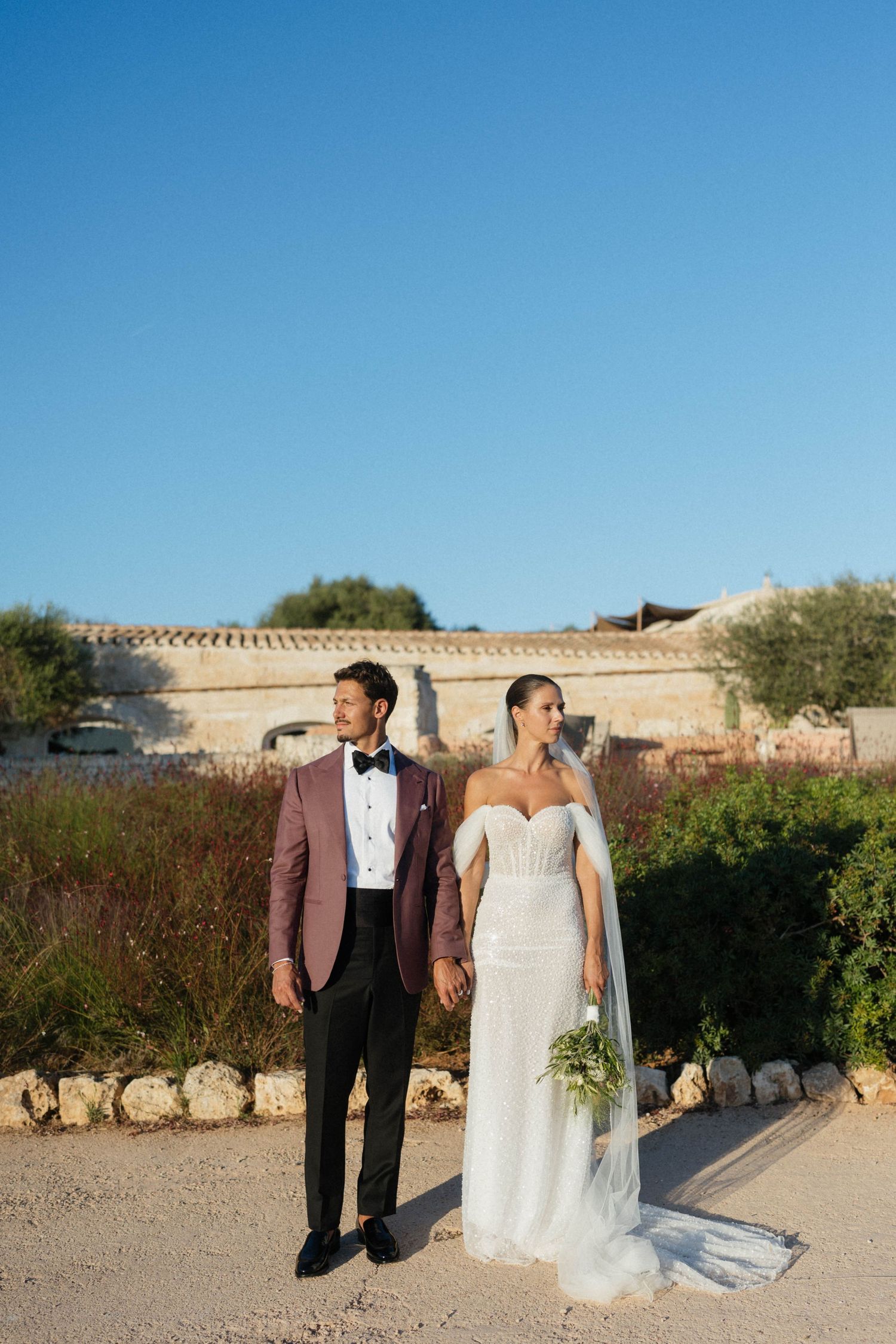 Smiling couple holding hands outdoors, the groom in a burgundy blazer and the bride in a beaded gown holding a green bouquet.