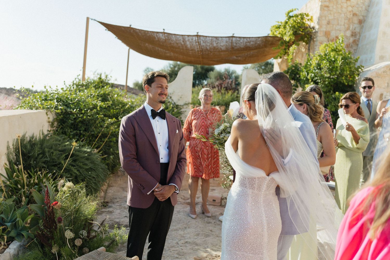 A groom in a burgundy blazer and bow tie waits at the outdoor altar as the veiled bride embraces a guest beneath a rustic canopy.