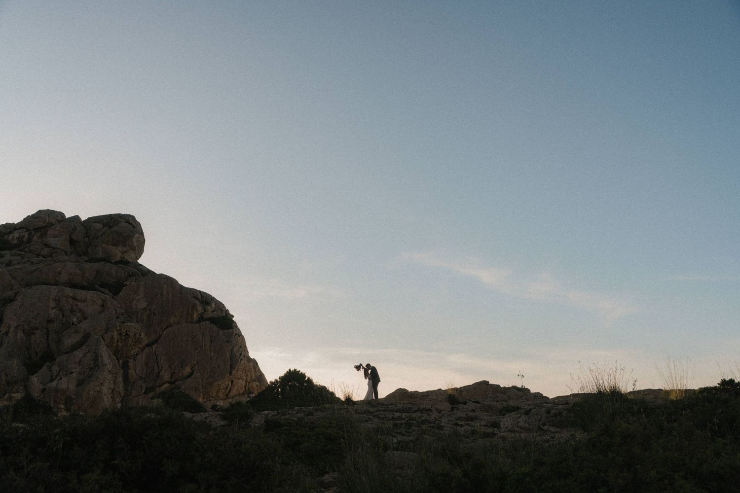 Silhouette of couple standing on rocky desert landscape at dusk with clear blue sky and large boulders.