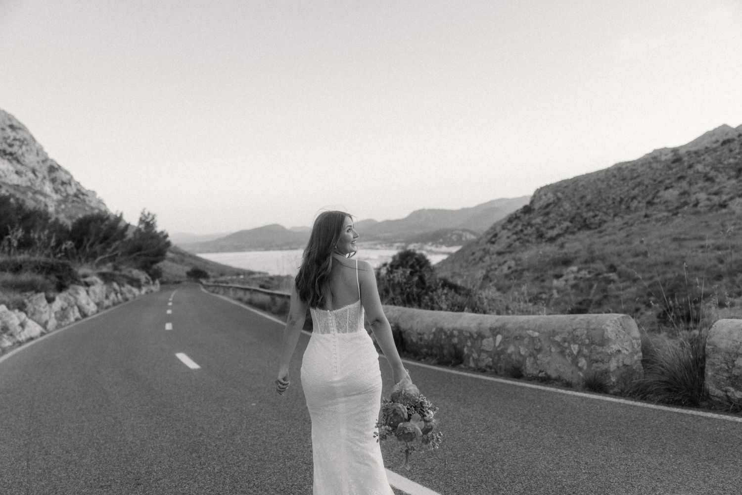 Bride in fitted white gown walks along scenic mountain road holding bouquet in elegant black and white photo.