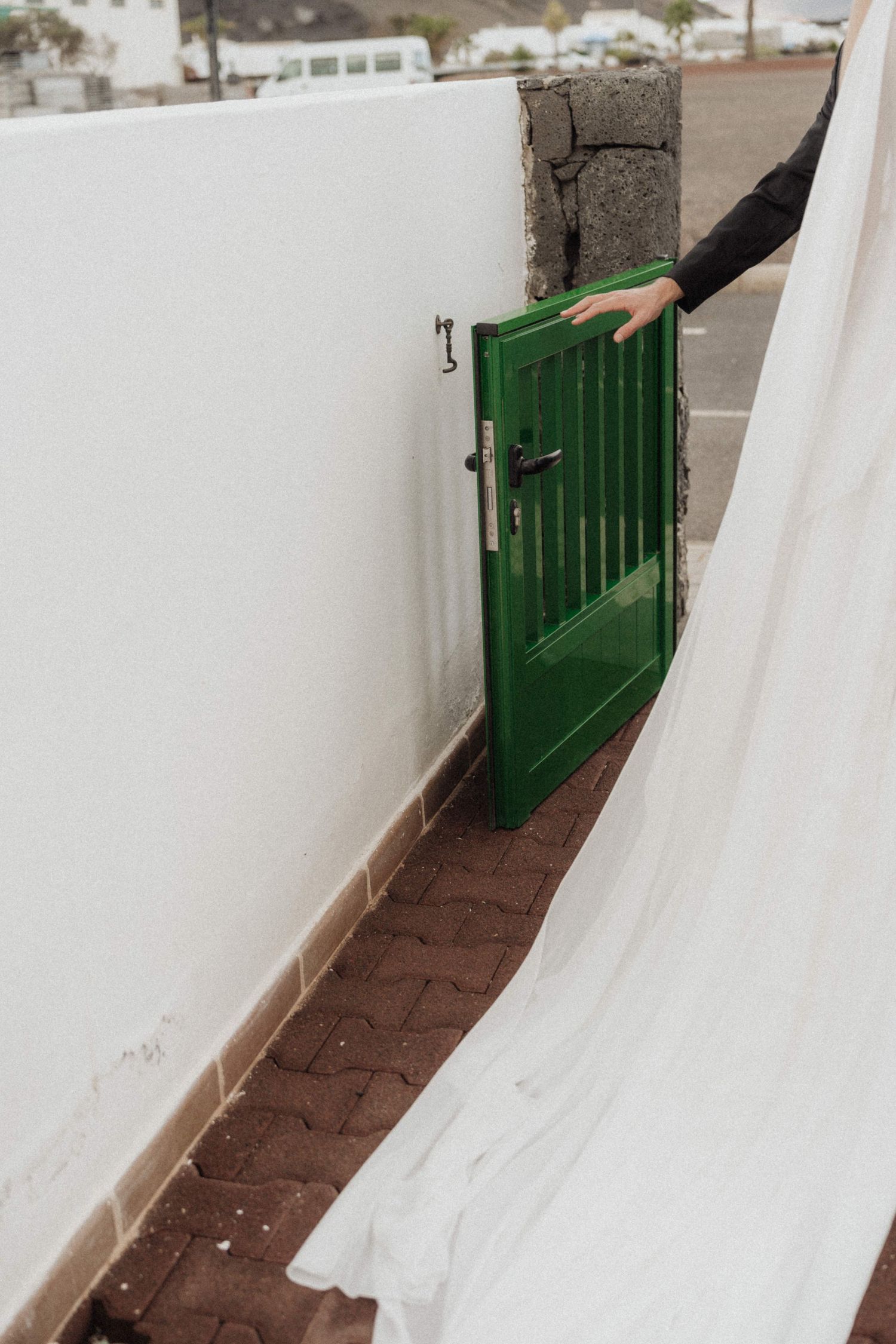 Hand opening green wooden gate in white wall leading to brick pathway outside.