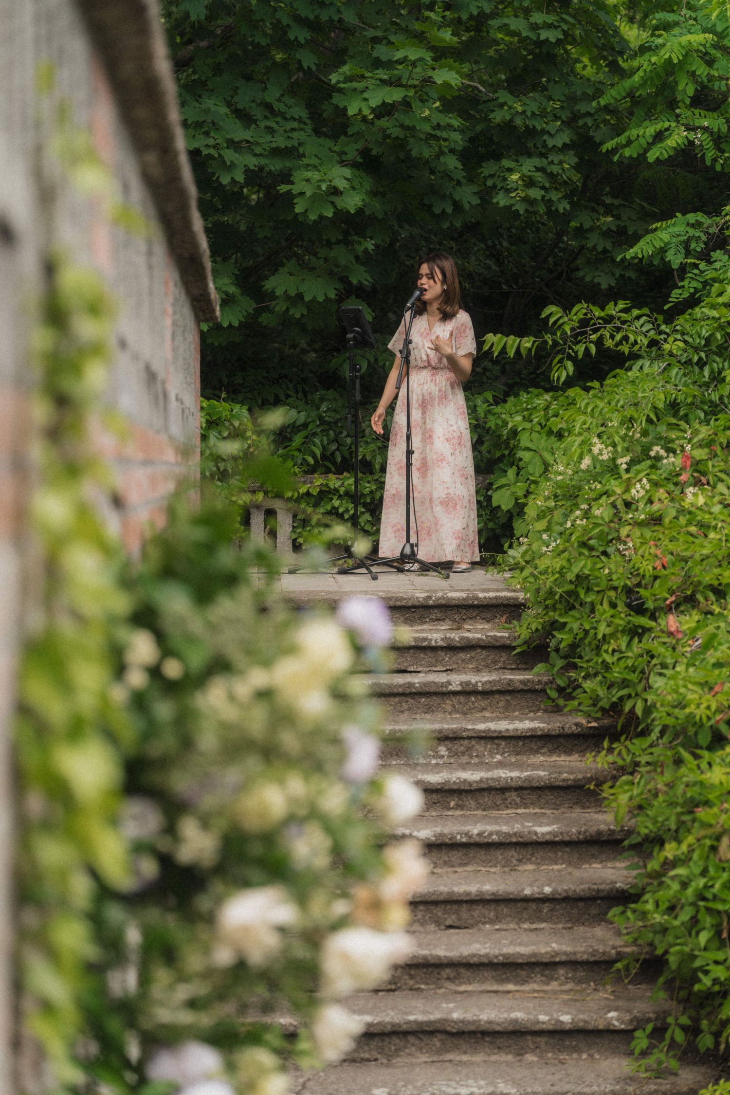 Woman in floral dress standing on stone garden steps surrounded by lush green foliage and white flowers.