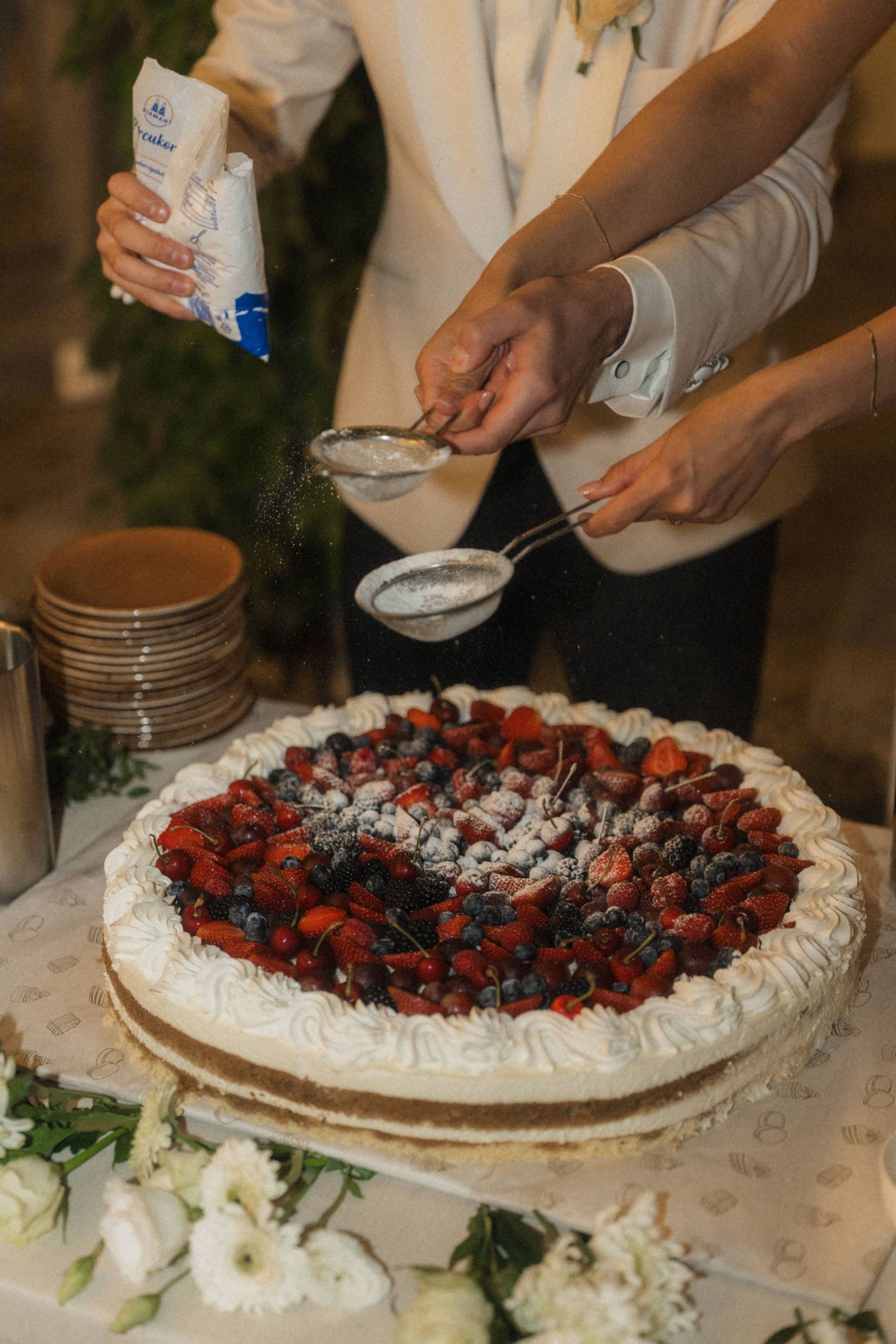 Couple dusting powdered sugar over a large wedding cake topped with fresh mixed berries.