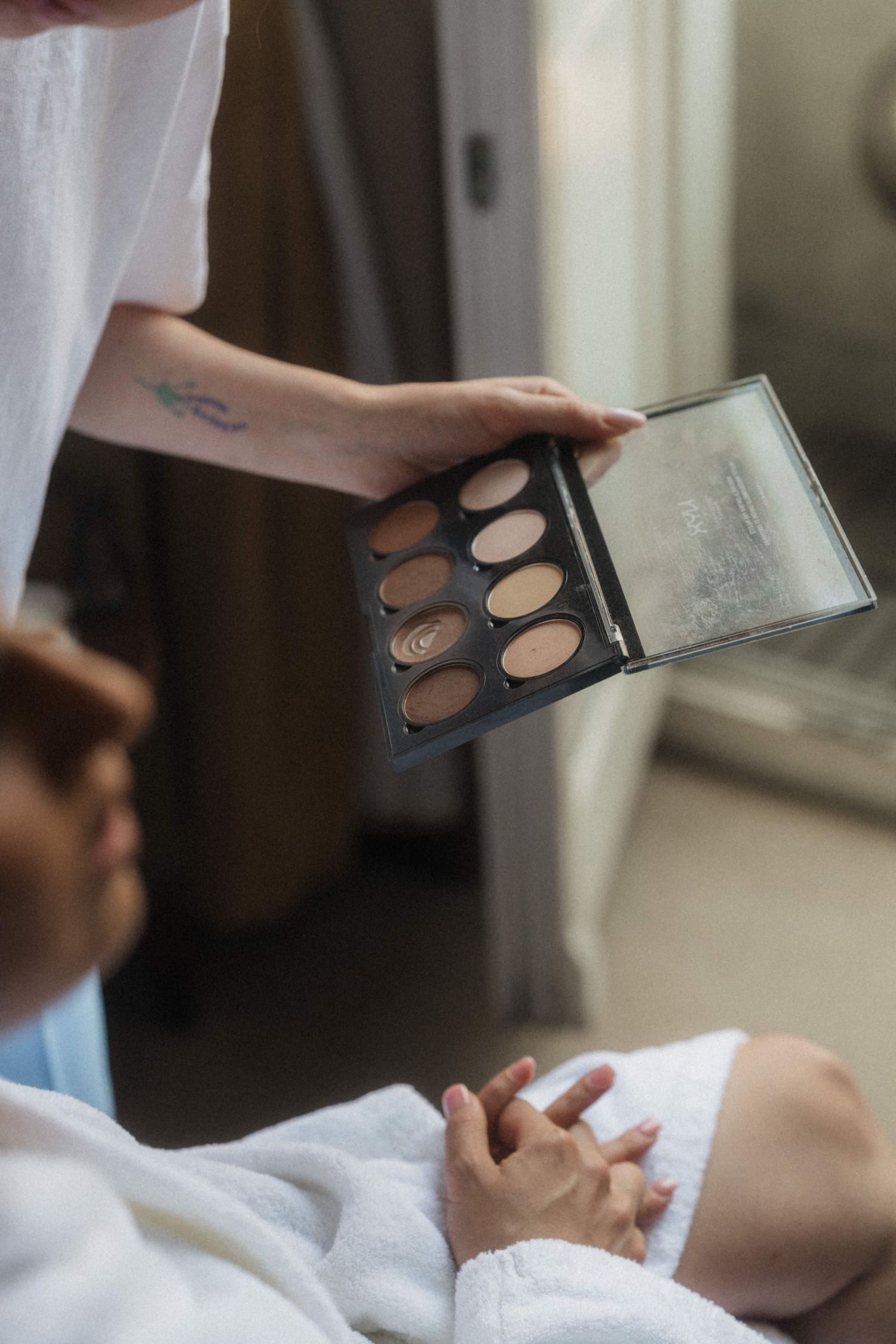 Makeup artist holding eyeshadow palette during bridal preparation in white robes.