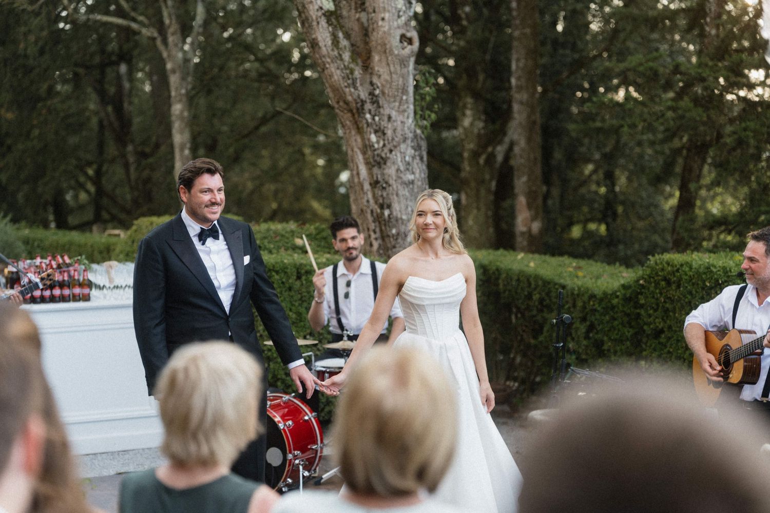Bride and groom smiling joyfully at outdoor wedding ceremony with live band performing in background.