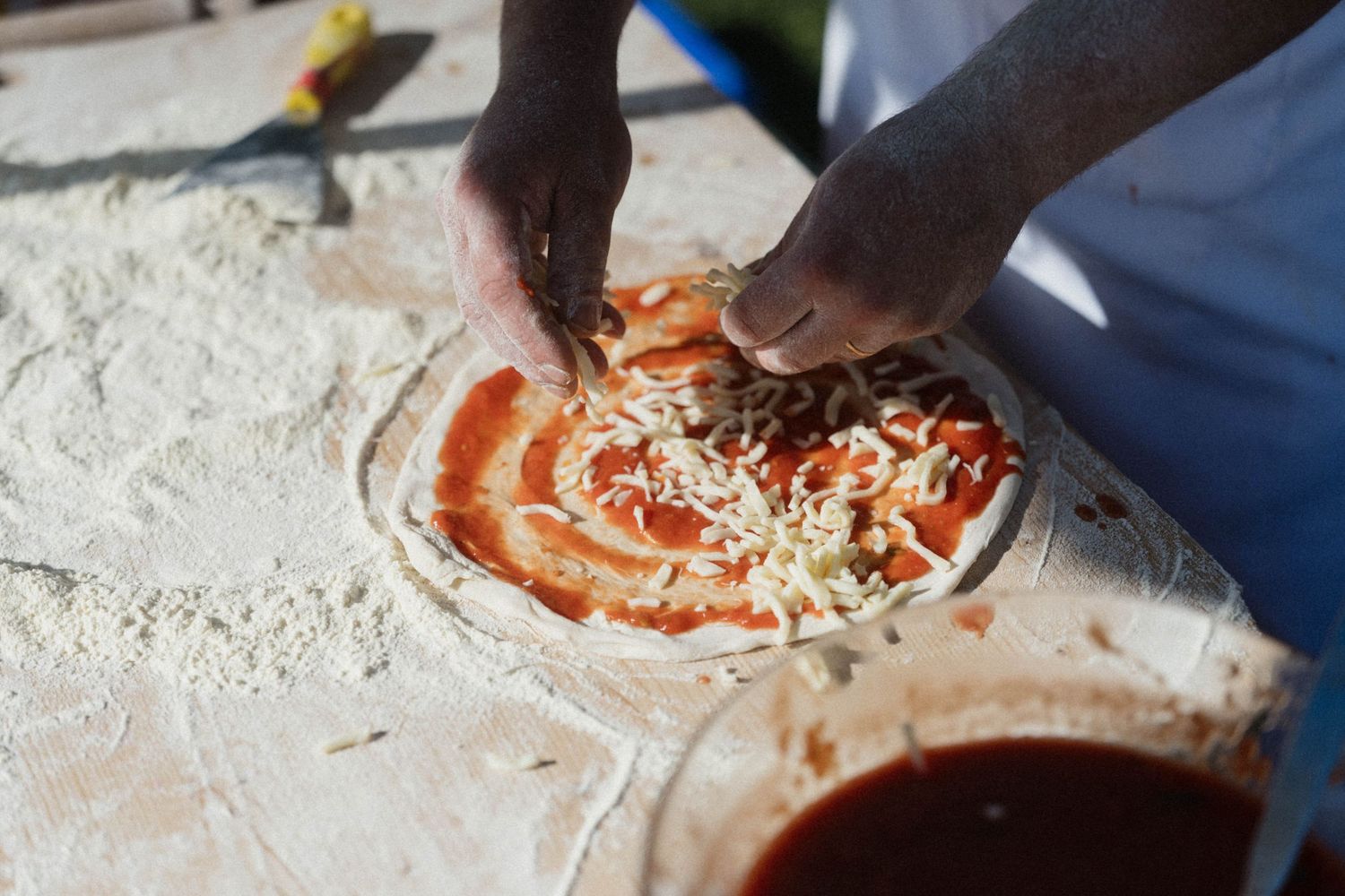 Chef topping a fresh pizza with cheese and tomato sauce on a flour-dusted surface.