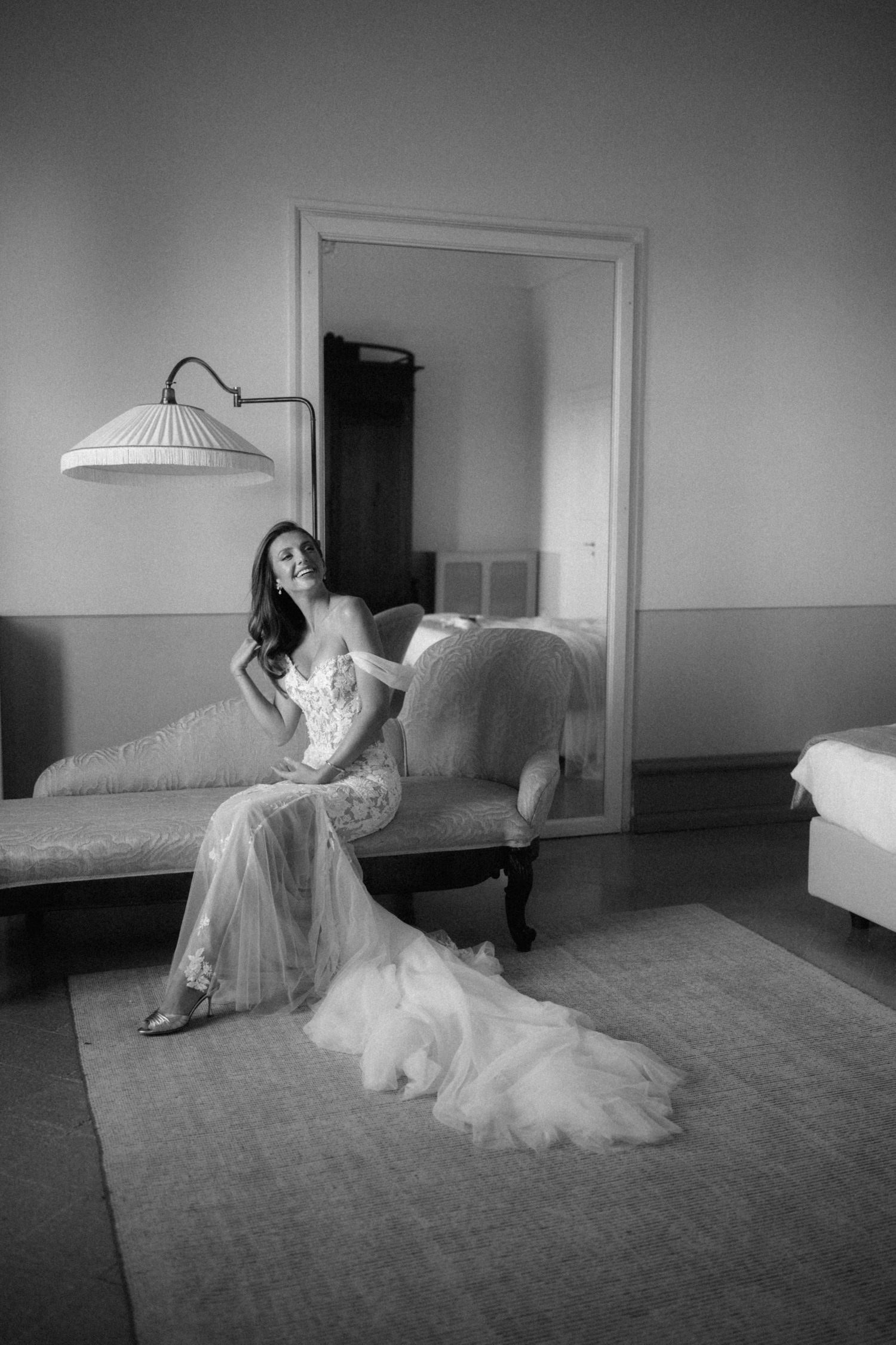 Black and white photo of smiling bride in lace gown seated on vintage chaise lounge.