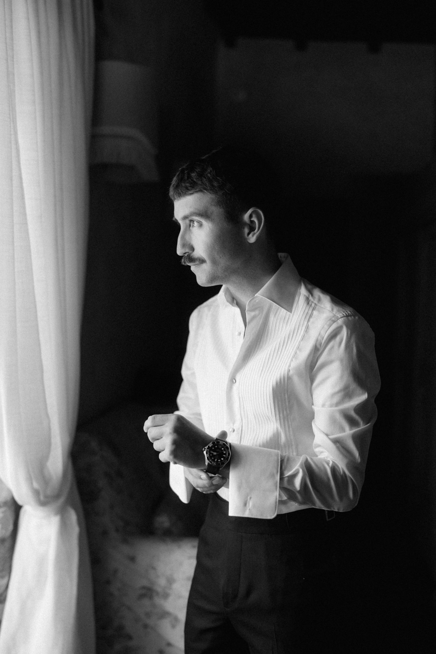 Elegant groom in white dress shirt and watch adjusting cufflinks by a window in black and white photo.