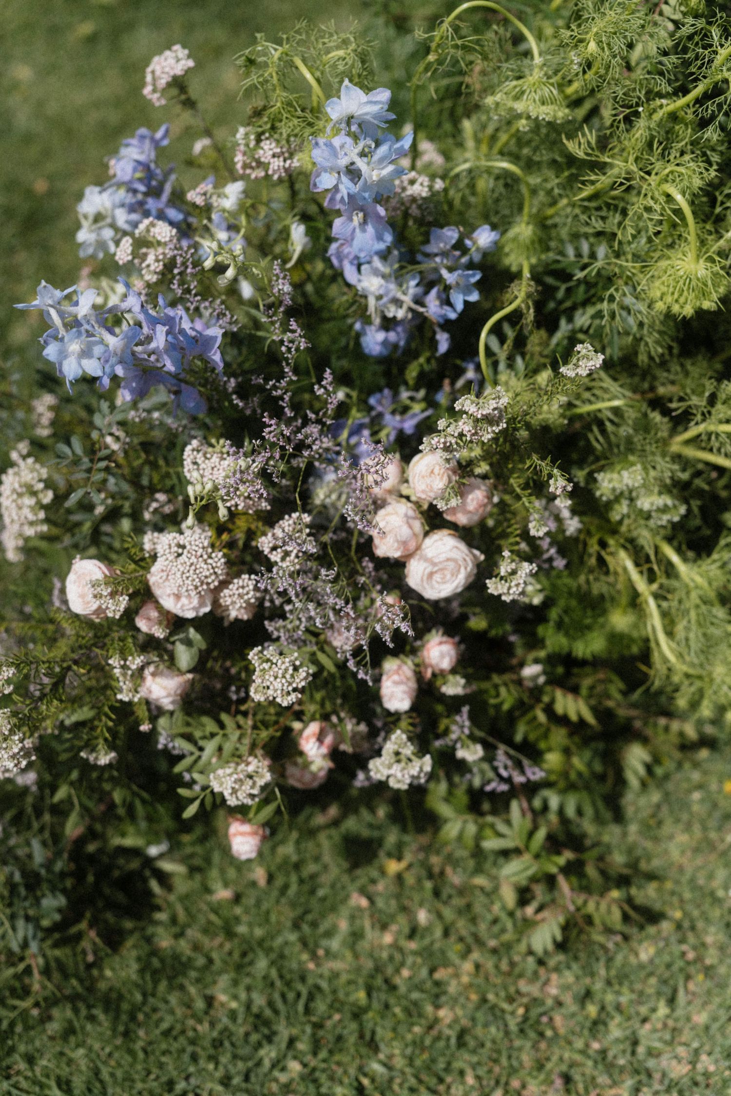 Rustic wedding bouquet with blue delphiniums, blush roses, white wildflowers, and lush greenery on grass.