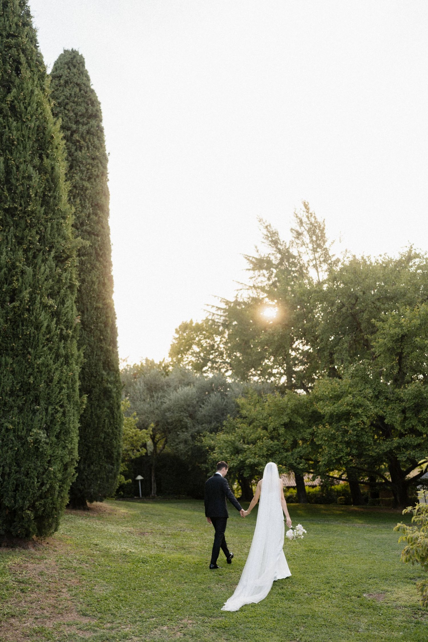 Bride and groom walking hand in hand through a lush garden at sunset, with tall cypress trees and warm golden light.