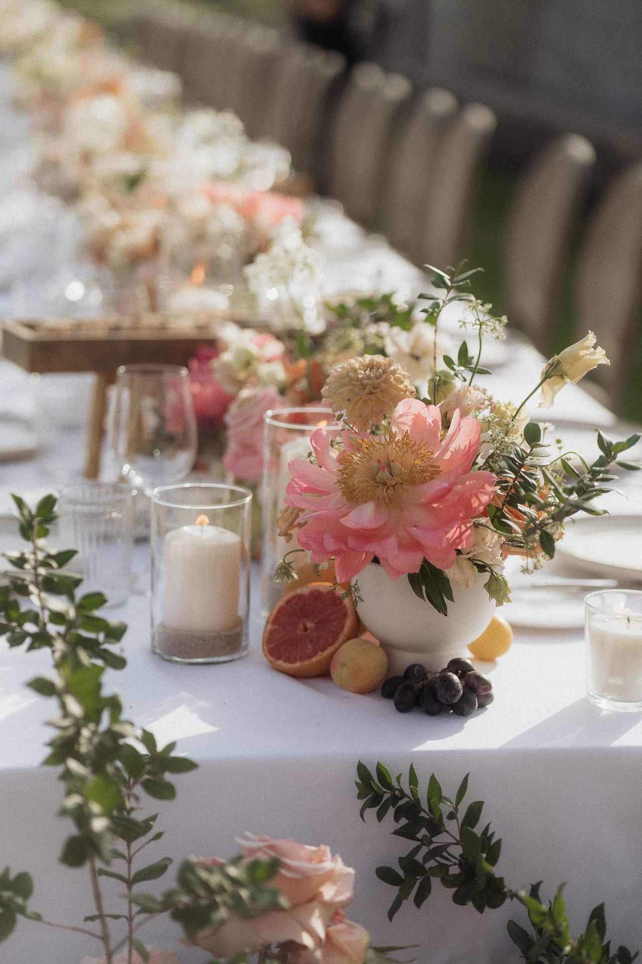 Elegant outdoor wedding table with pink floral centerpieces, candles, and fresh fruit on white linen.