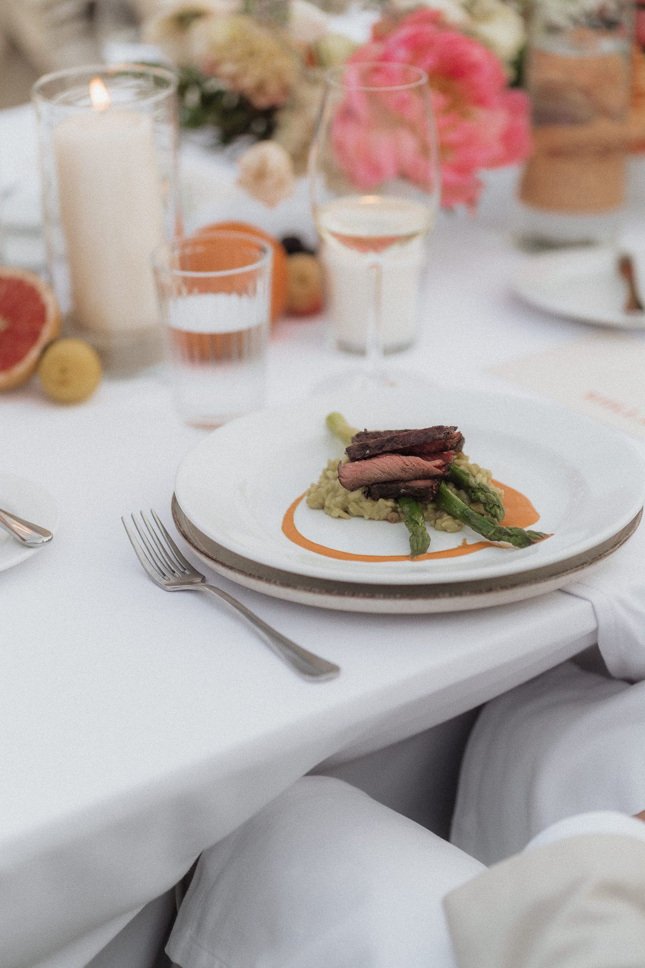 Elegant wedding dinner plate with seared meat, asparagus, and sauce on white tablecloth with candles and flowers.