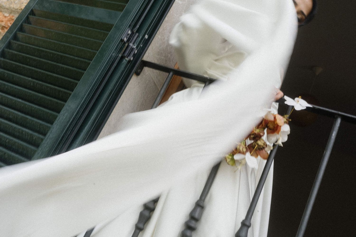 Bride in white wedding dress with flowing veil caught by wind on elegant staircase railing.