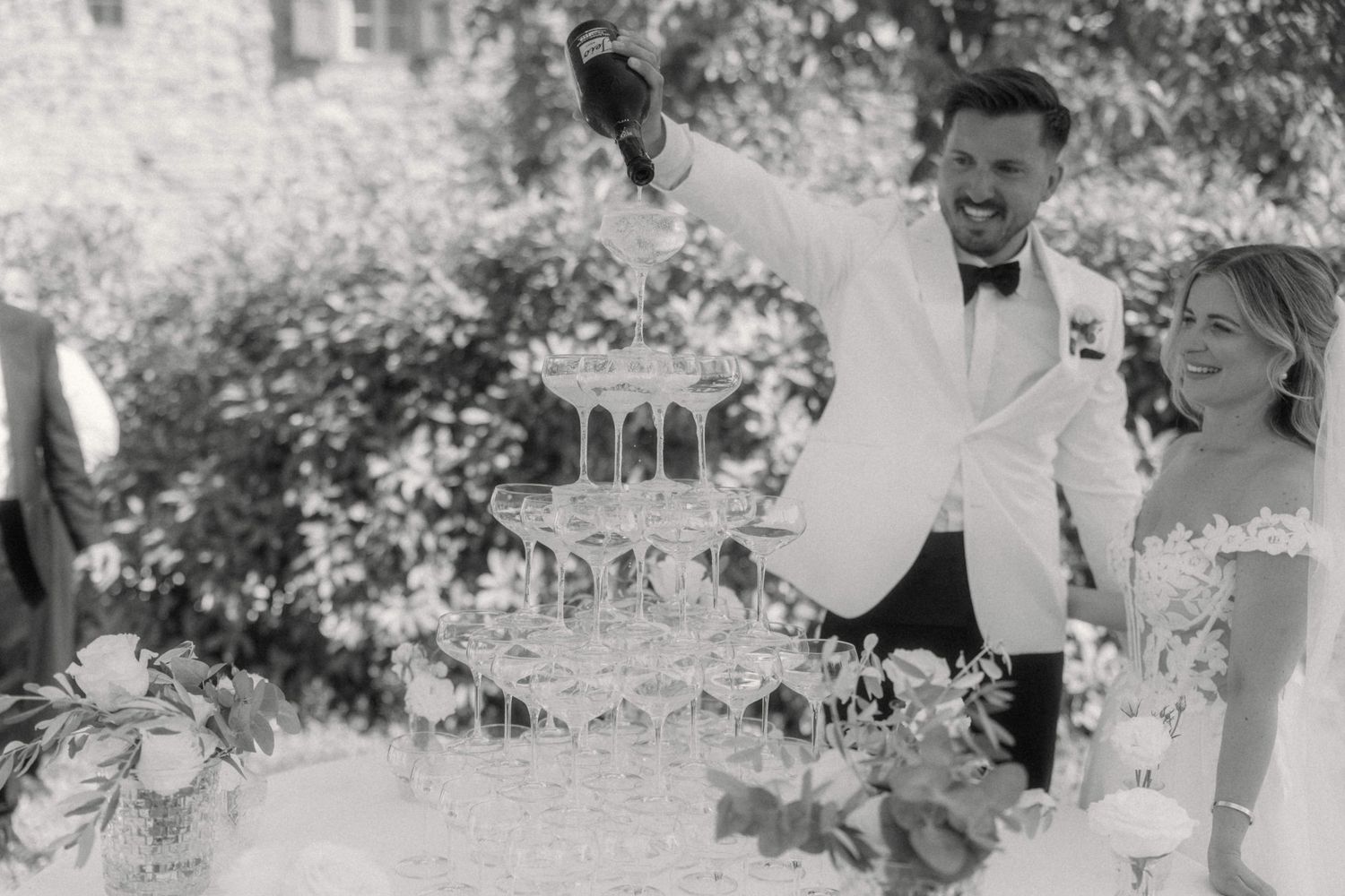 Groom in white tuxedo pouring champagne tower at elegant black and white wedding reception.
