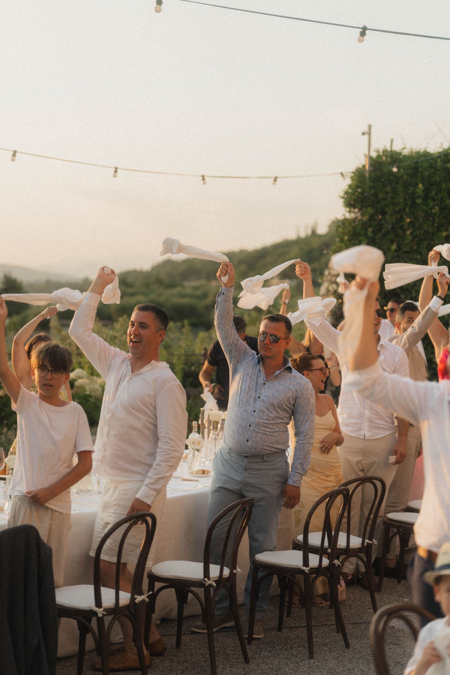 Joyful wedding guests waving white napkins at outdoor reception dinner under string lights at sunset.