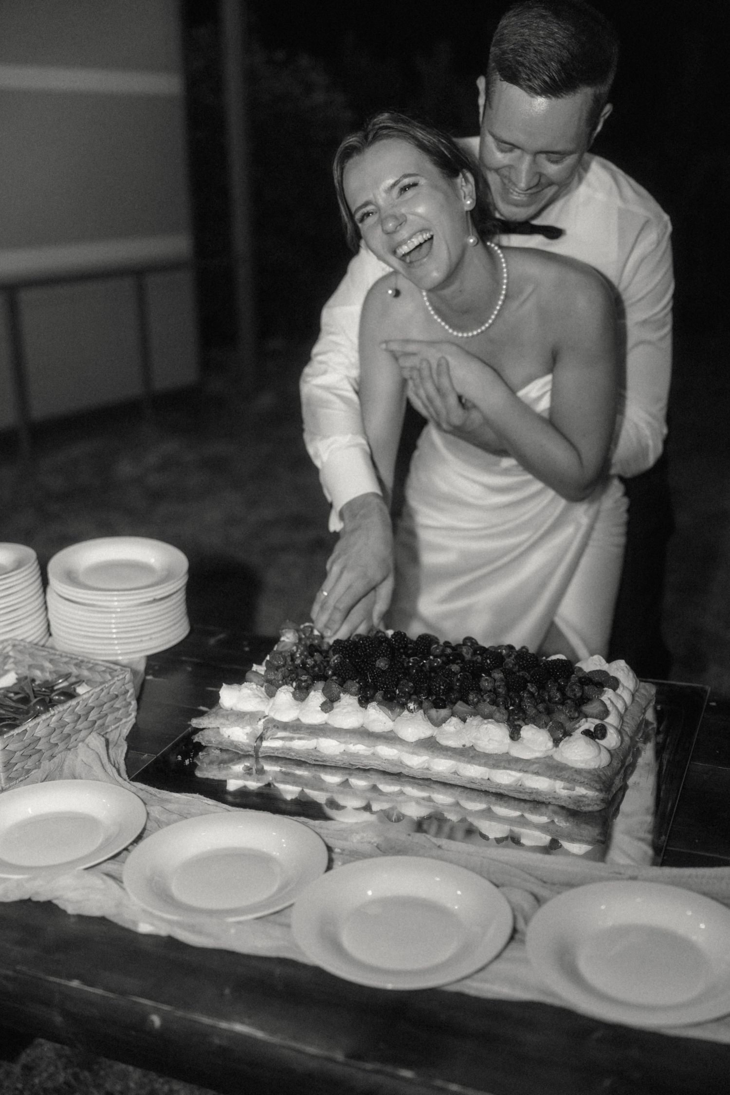 Black and white photo of laughing bride and groom cutting layered fruit wedding cake together.
