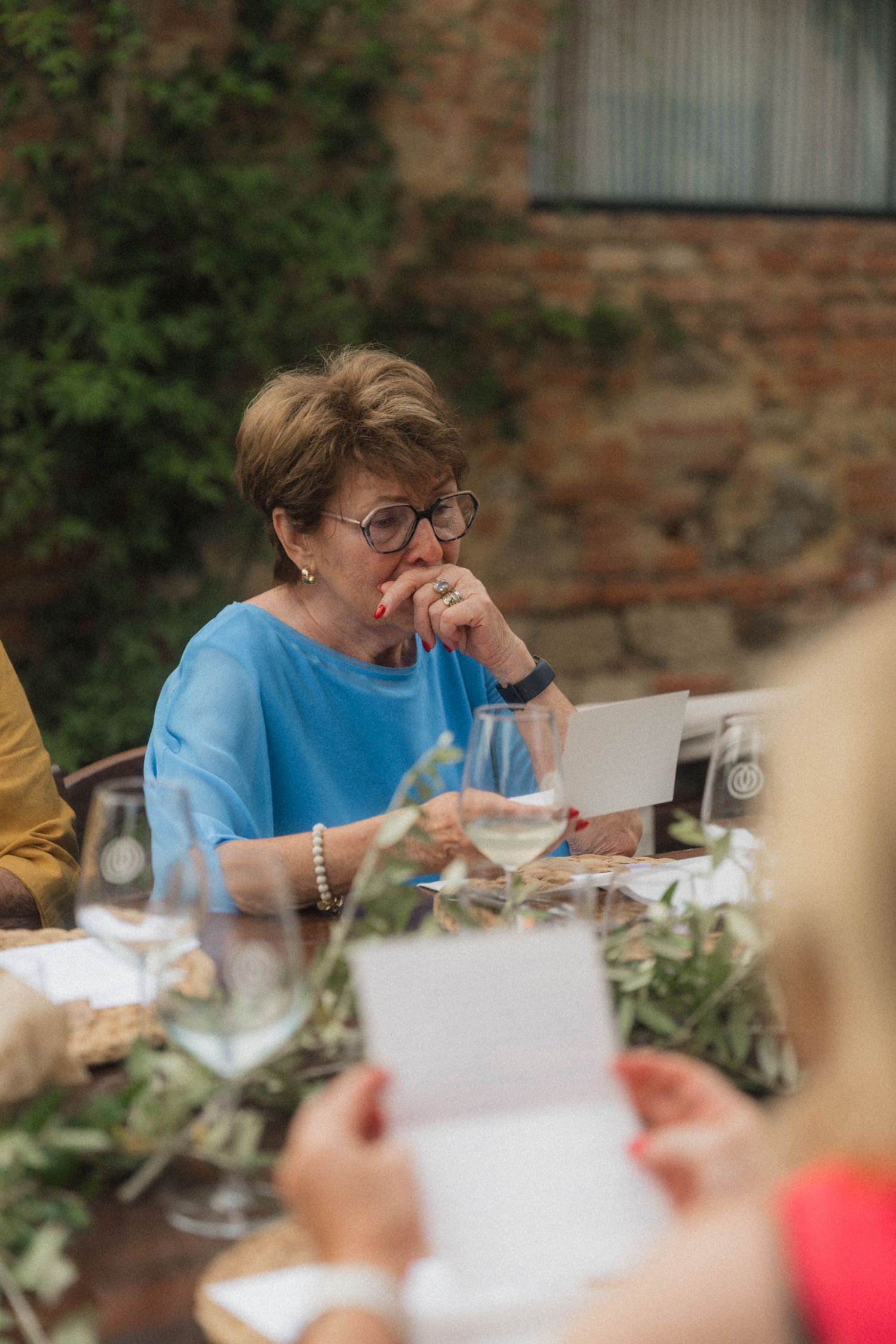 Emotional female wedding guest in blue blouse reading heartfelt speech card at outdoor reception table.
