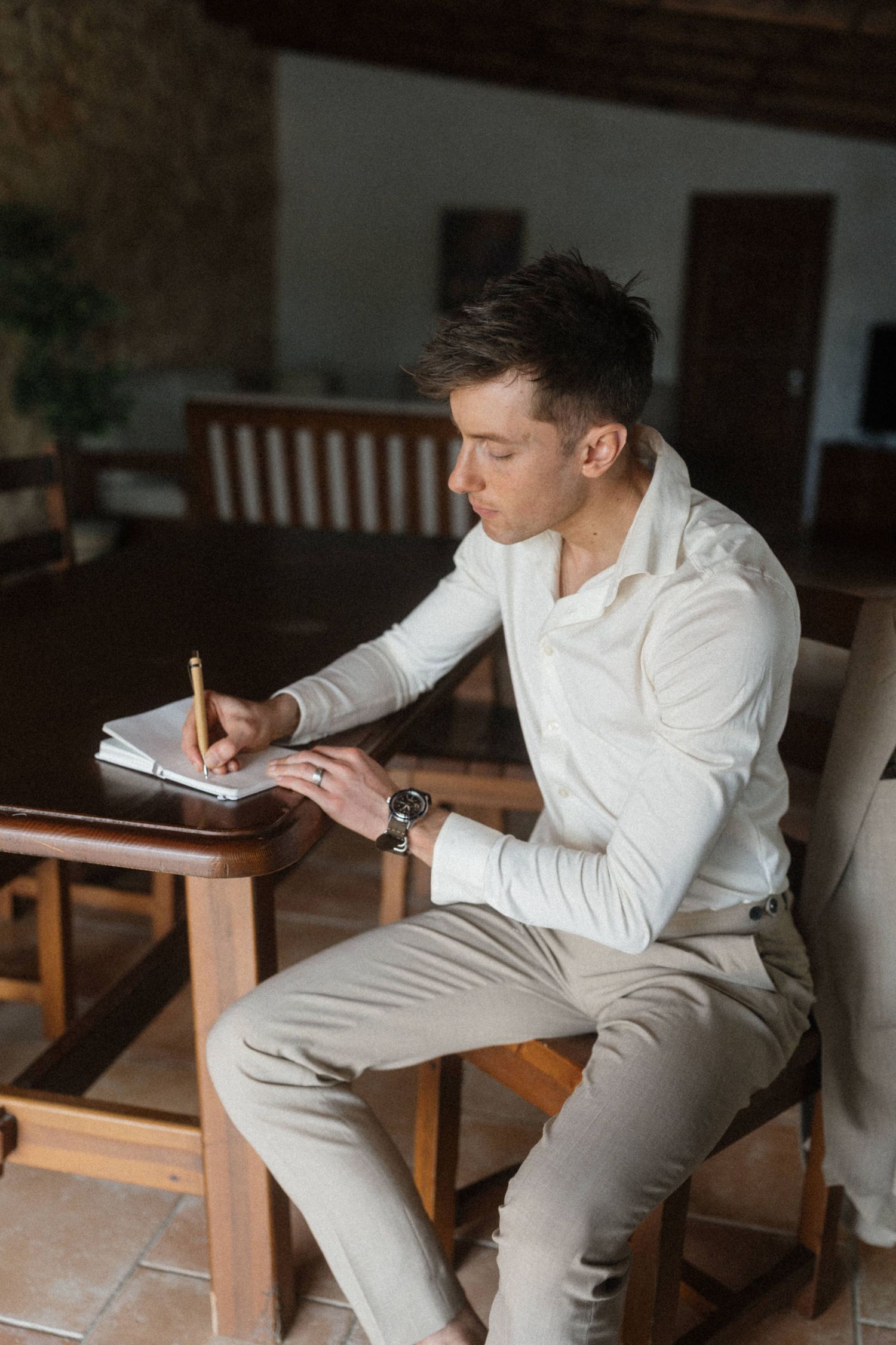 Young man in white shirt writing in notebook at wooden table, focused and wearing a watch.