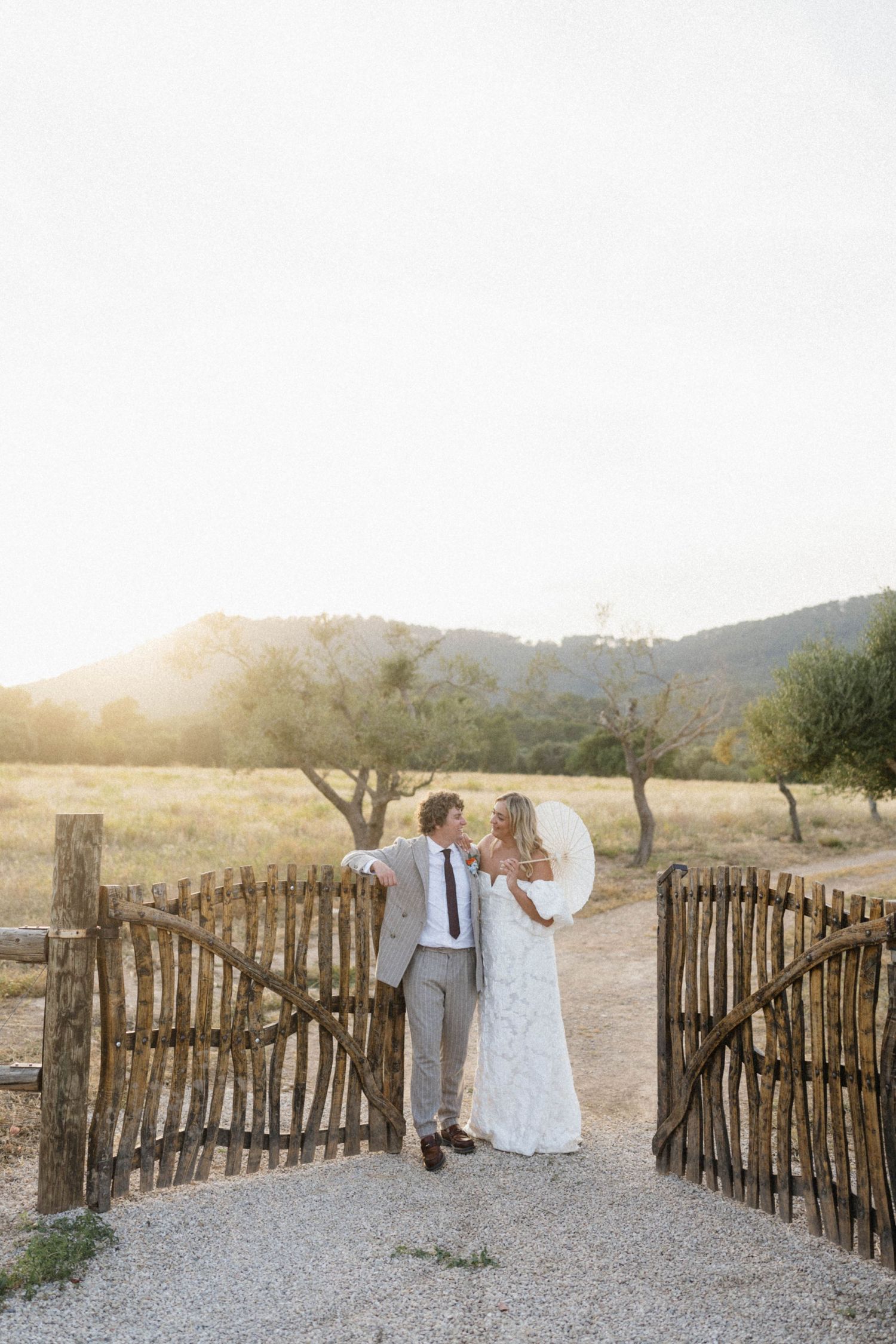 Bride and groom embracing at rustic wooden gate during golden hour sunset in scenic countryside.