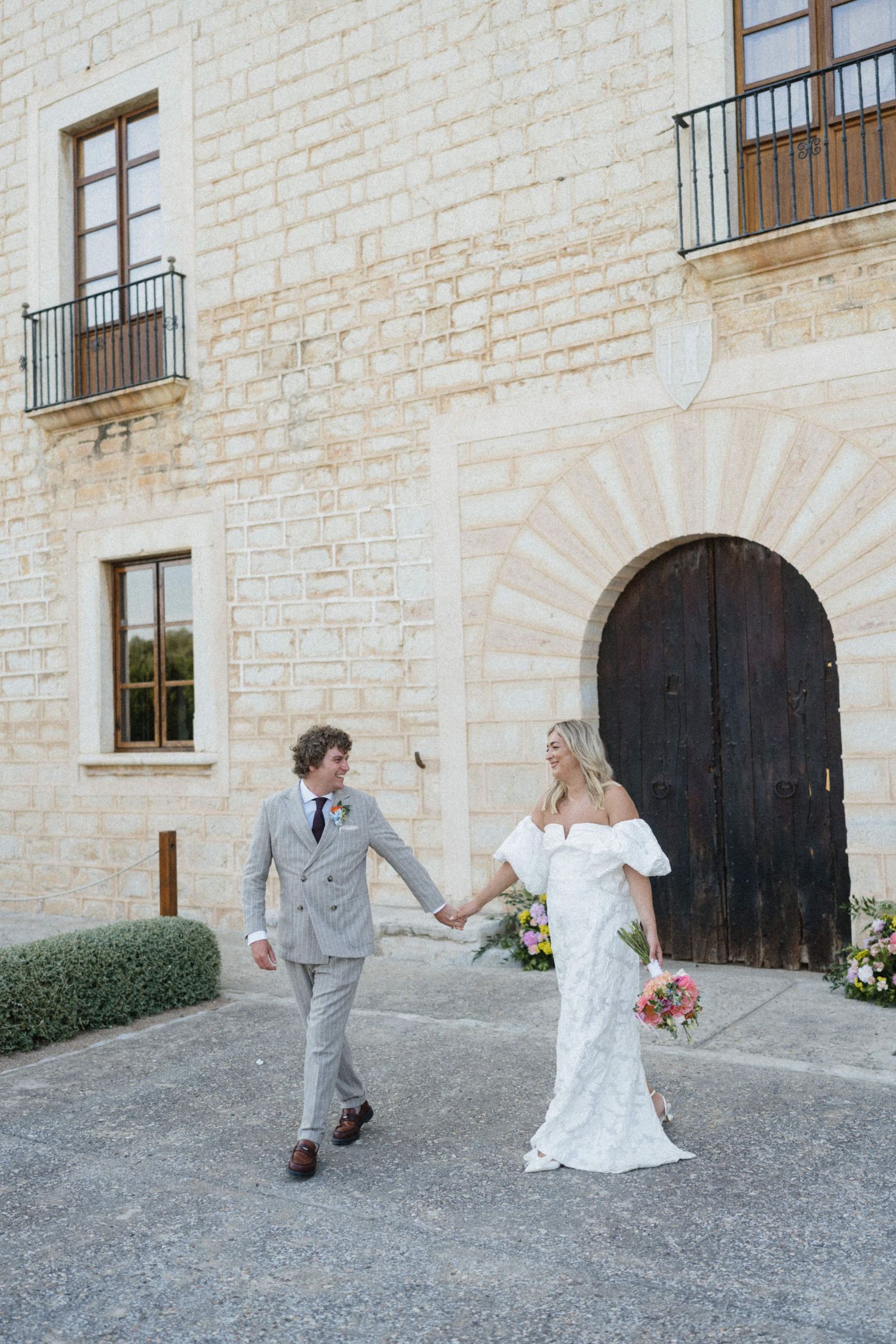 Bride and groom hold hands walking in front of a rustic stone building with arched wooden door.