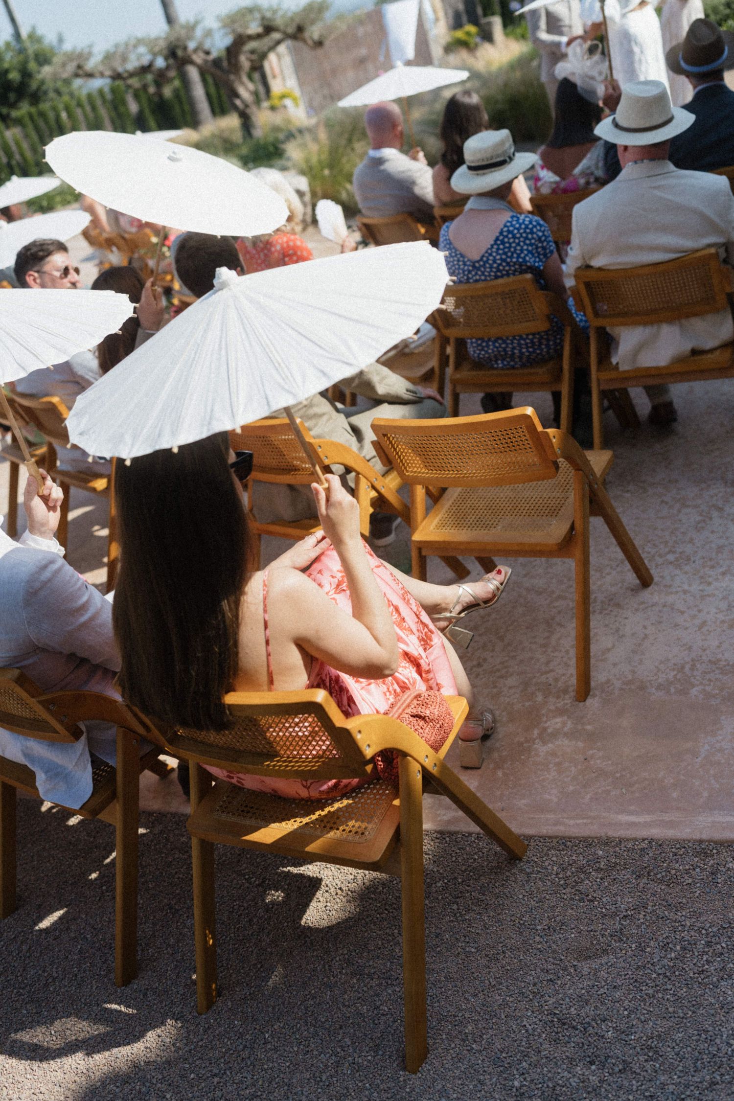 Guests seated in wooden chairs holding white parasols at an outdoor wedding ceremony on a sunny day.