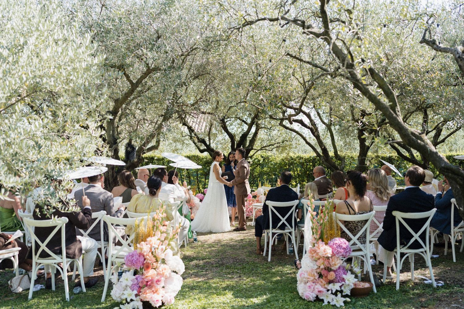Romantic outdoor wedding ceremony under olive trees with guests seated in white chairs.