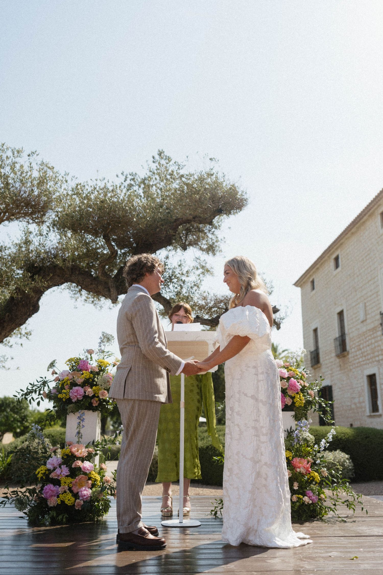 Couple exchanging vows at outdoor wedding ceremony with floral arrangements and olive tree backdrop.