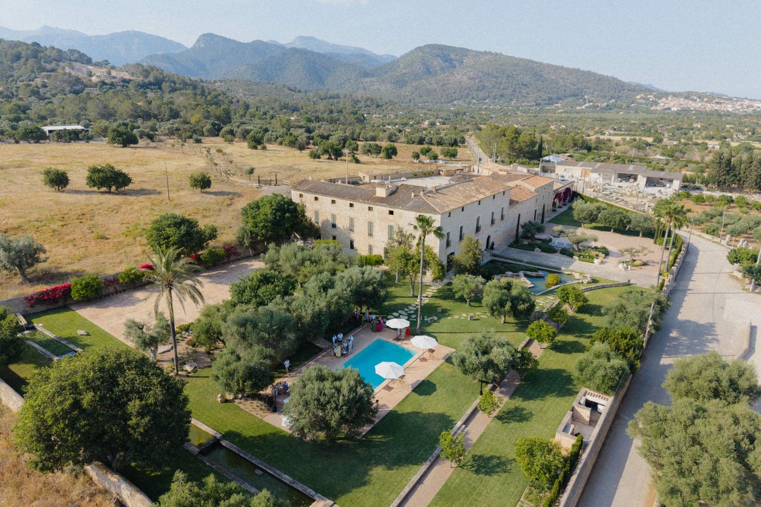 Aerial view of a large rustic estate with pool, lush gardens, and mountain backdrop.