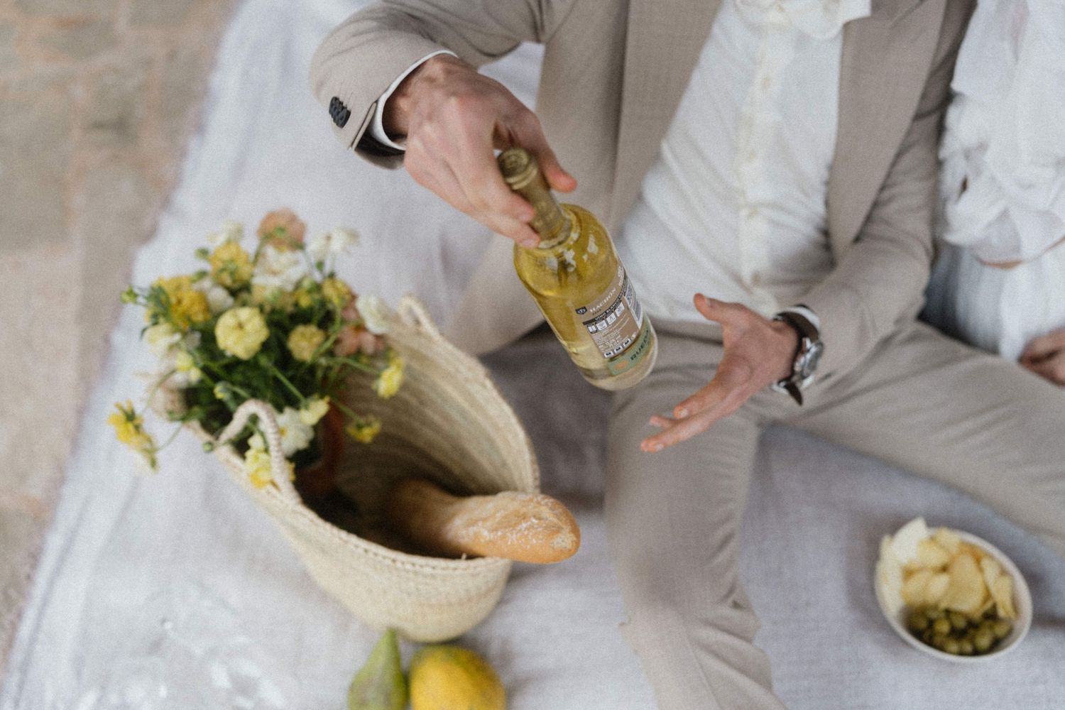 Man in beige suit opening a bottle at a picnic with bread, flowers, and fruit on a white blanket.